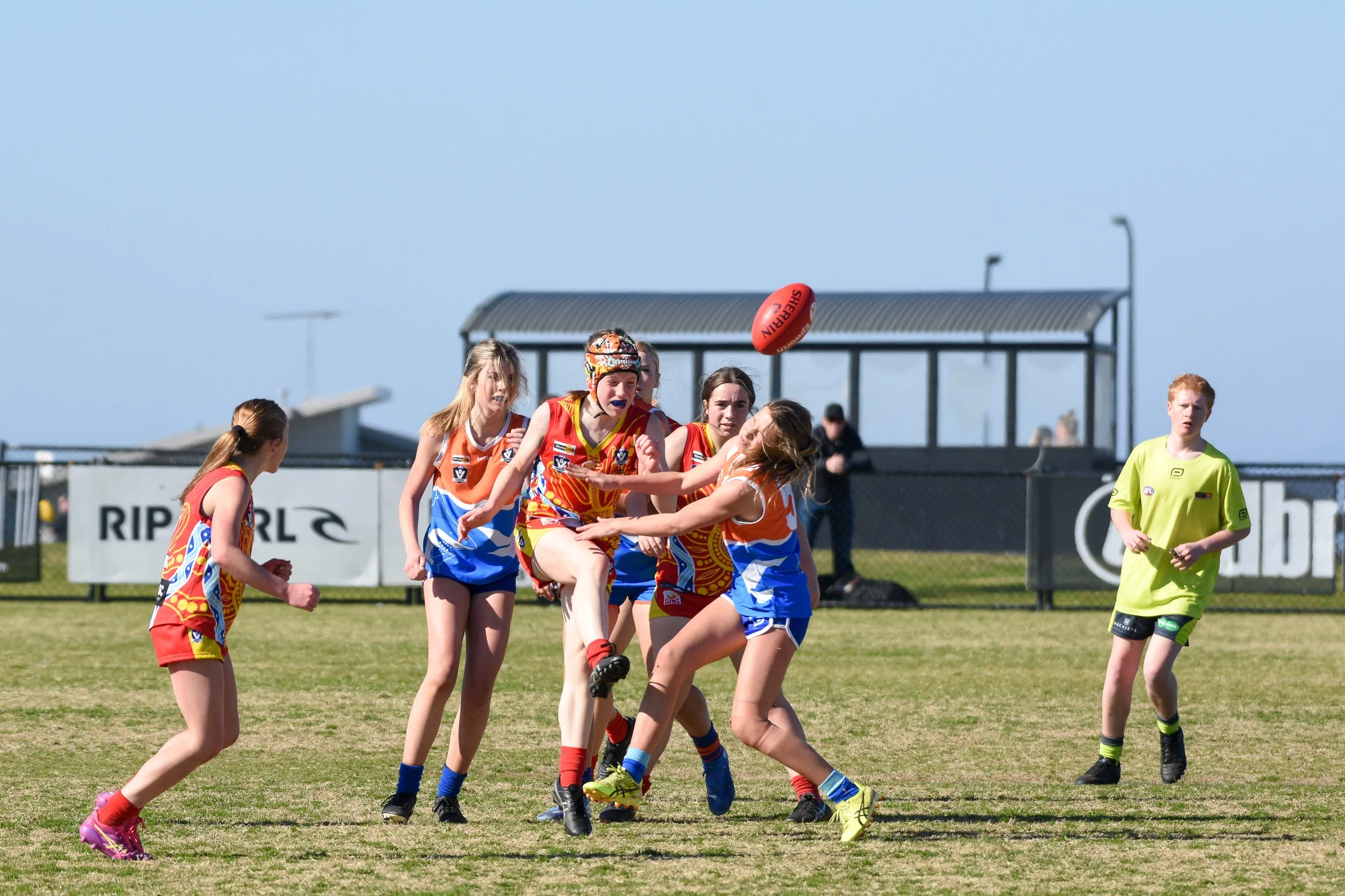 A group of young women playing Australian rules football on a grassy field during daytime, with one player jumping to catch the ball while others attempt to block or defend, and a referee observing nearby.