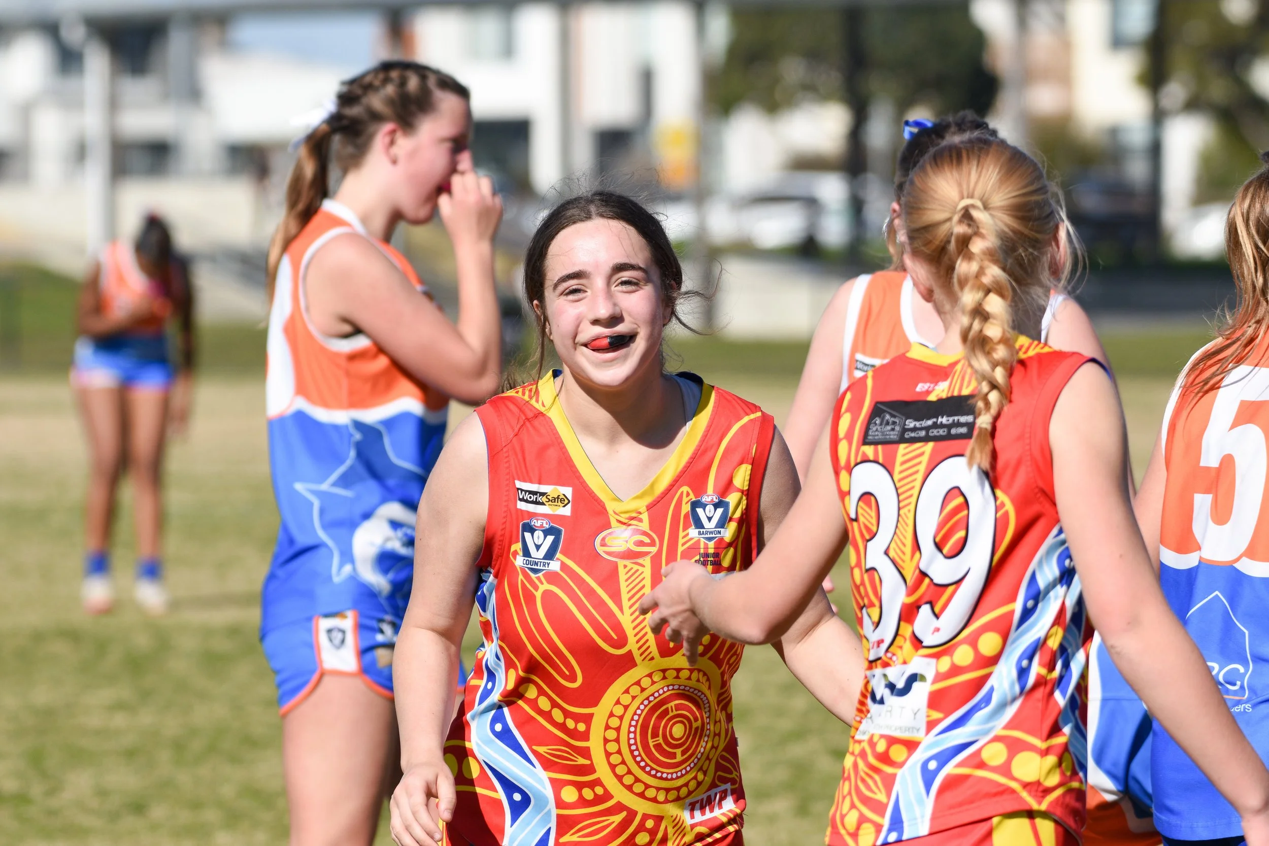 Group of young female Australian rules football players on the field, some are smiling and interacting after a game.