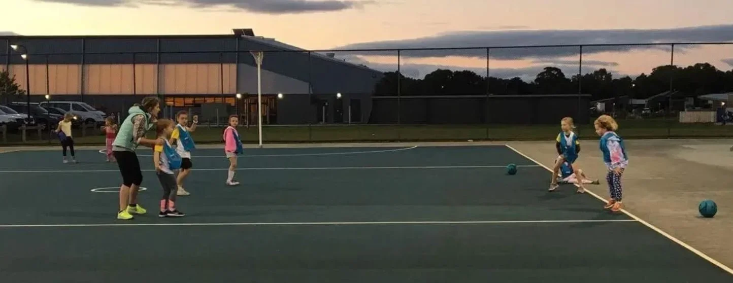 Children participating in a soccer drill on a court at dusk with coach, with balls and a large building in the background.