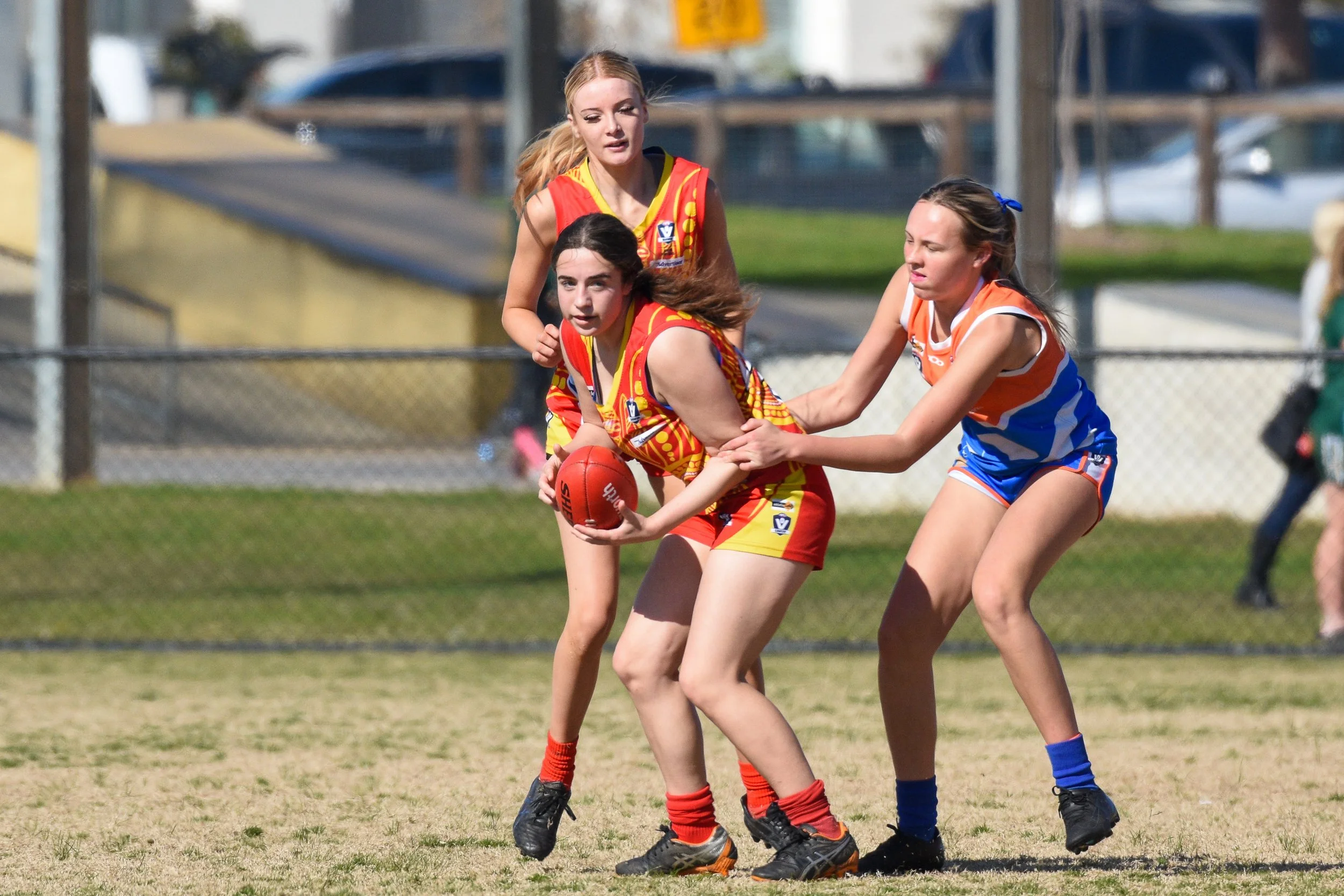 Women playing Australian rules football on a grassy field, with one player holding a red football while two others attempt to block or tackle her, all wearing colorful jerseys and shorts.