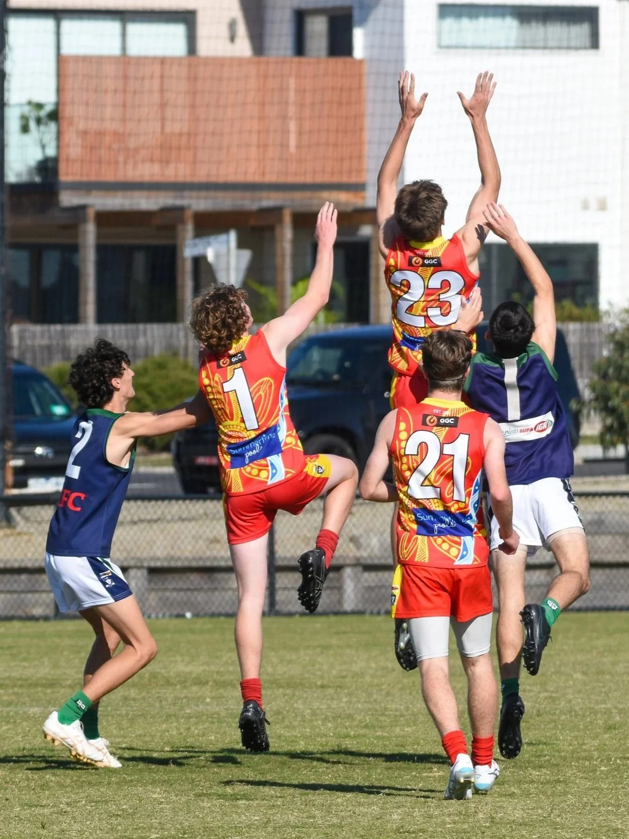 Children playing a game of footy on a grassy field, with some children jumping and reaching for the ball, wearing colorful sports uniforms.