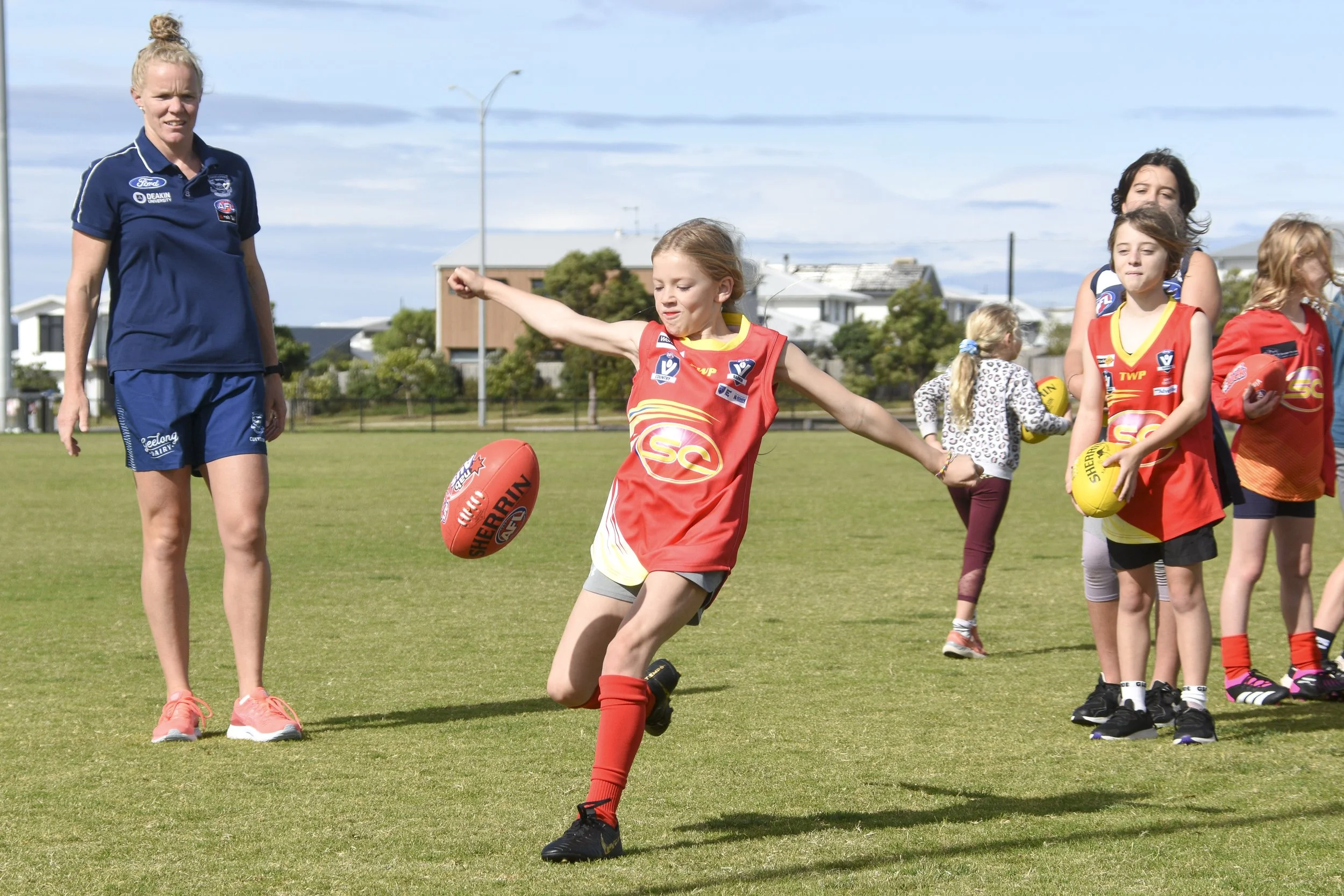 Children playing Australian Rules football on a grassy field, with an adult coach or supervisor observing. The children are wearing team jerseys and holding footballs.