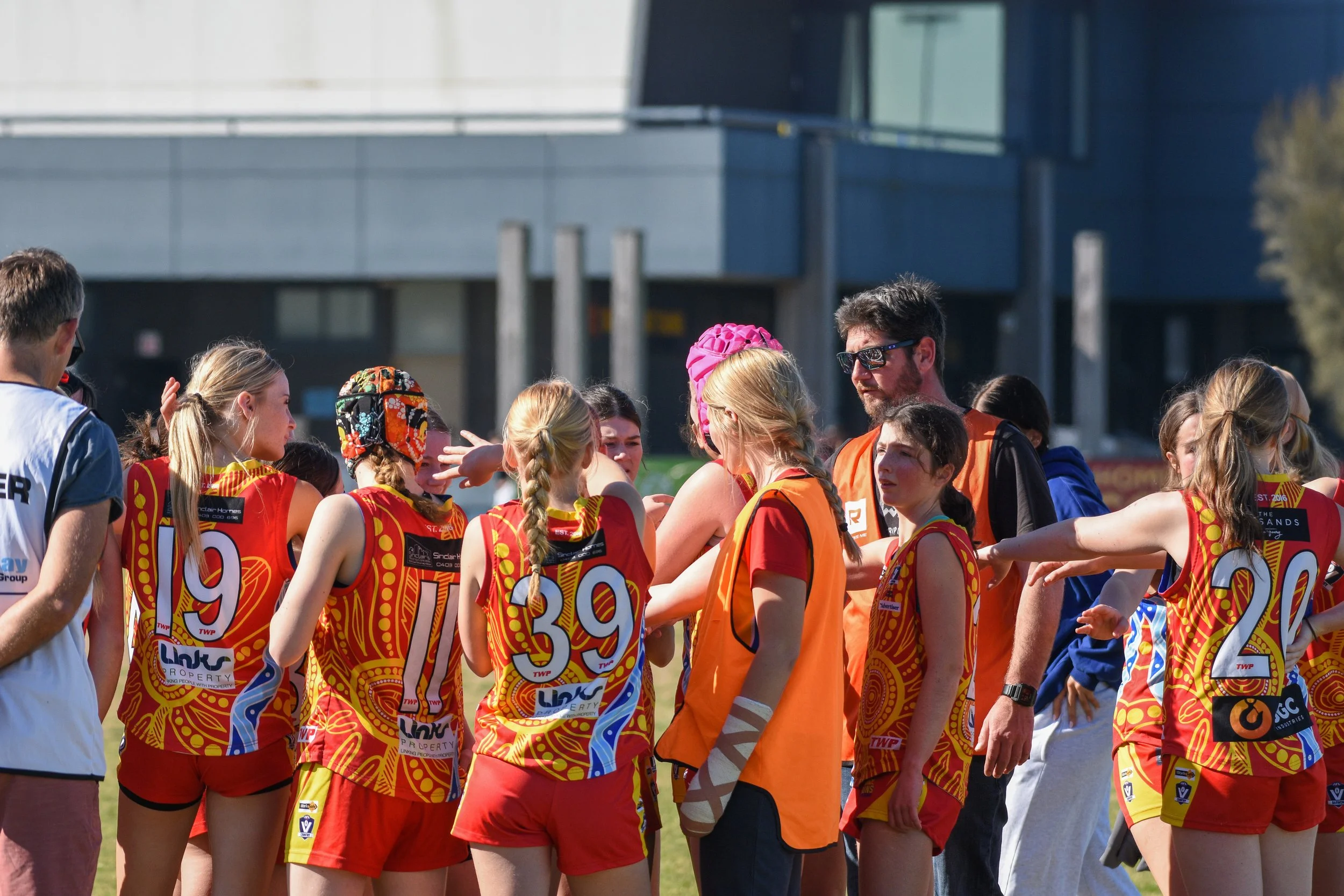 A group of female Australian rules football players in red and yellow uniforms are gathered in a huddle on the field with coaches, some wearing orange bibs, during daytime.