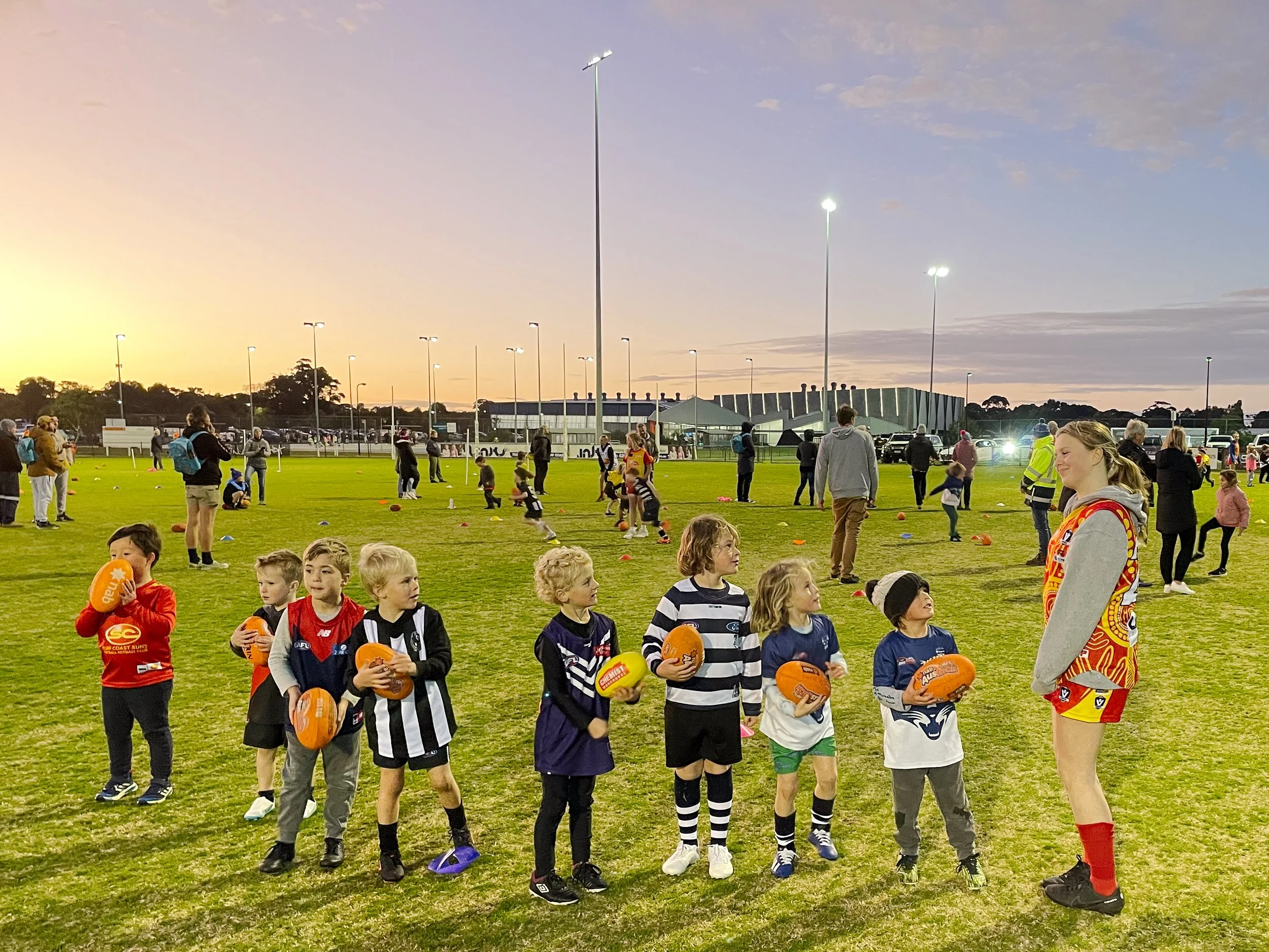 Group of young children with footballs standing in a line on a grassy field during evening, with women and others in the background, some practicing sports and others watching.
