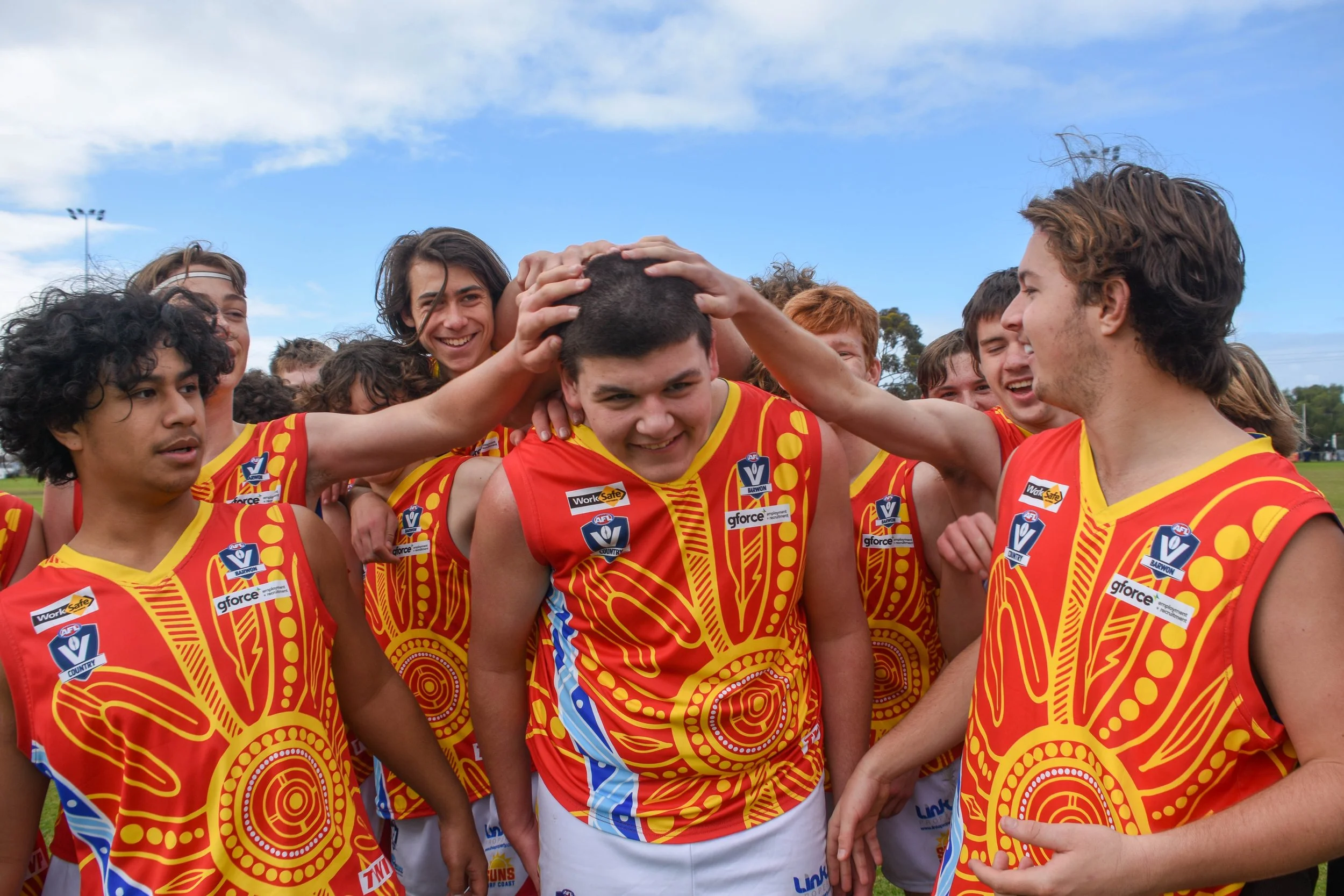 A group of young men in red and yellow sports jerseys celebrating on a field, with one person in the center smiling as others pat his head.