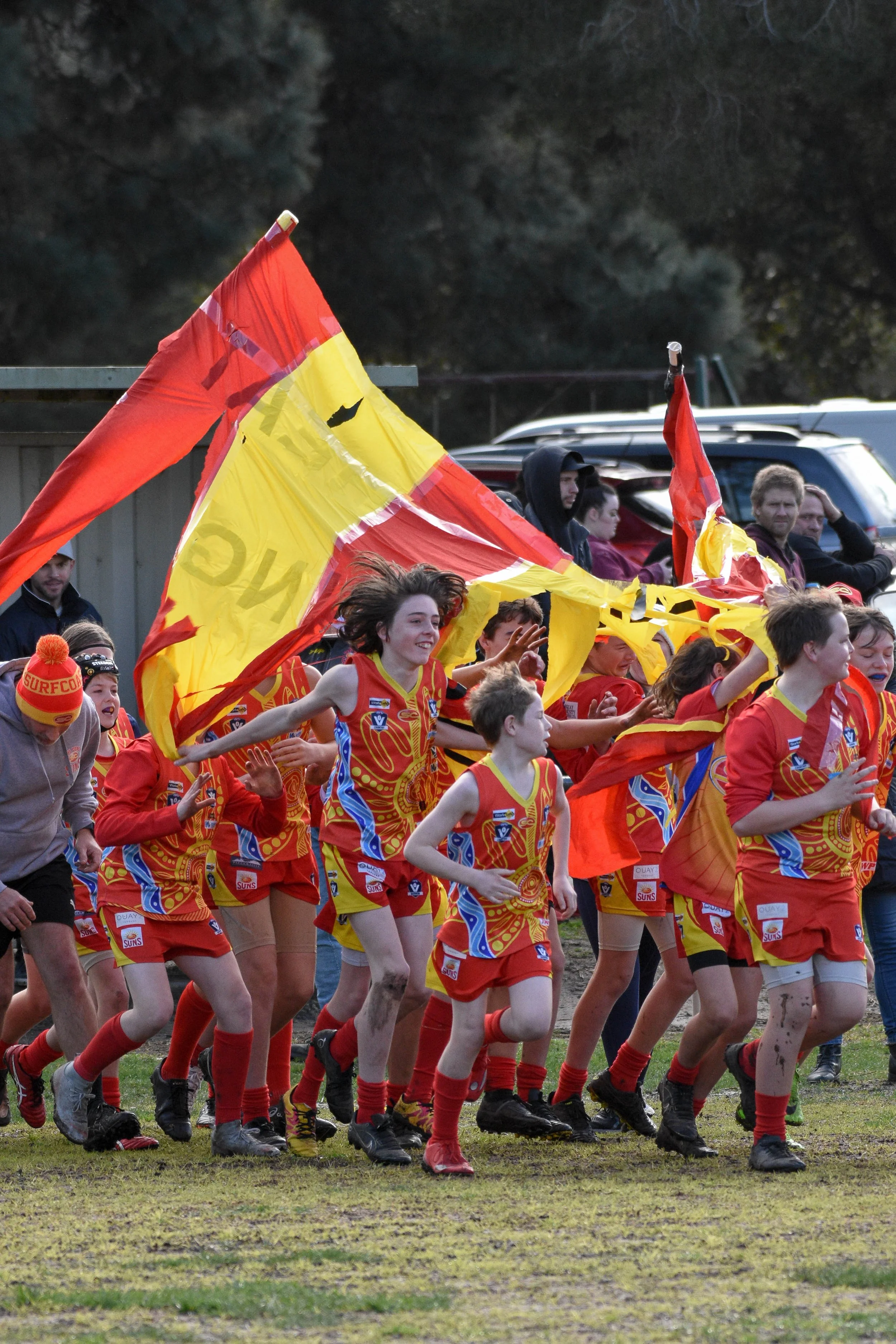 Young athletes in colorful uniforms running on a field during a sporting event, with spectators in the background, and a large flag being waved.