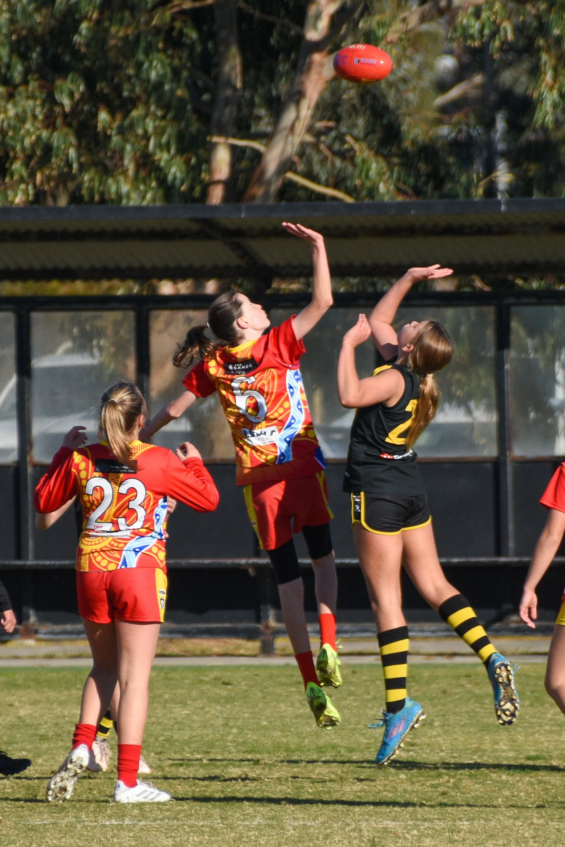 Women playing Australian rules football on a grassy field, with one player jumping to catch or hit the red ball in mid-air, dressed in black and yellow uniform, and two players in red and yellow uniforms.