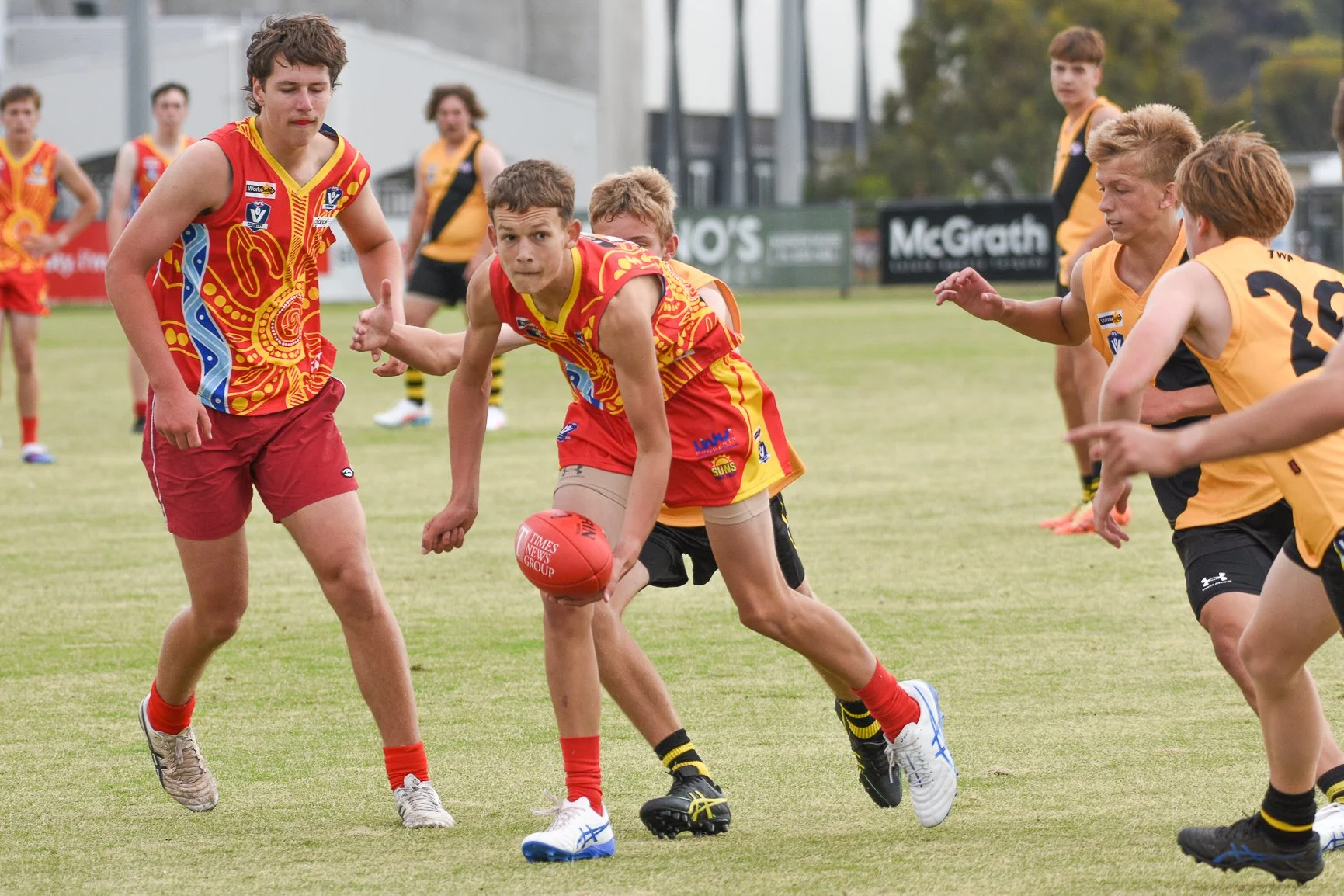 Australian rules football game with young players on a field, some running and others reaching for the ball.