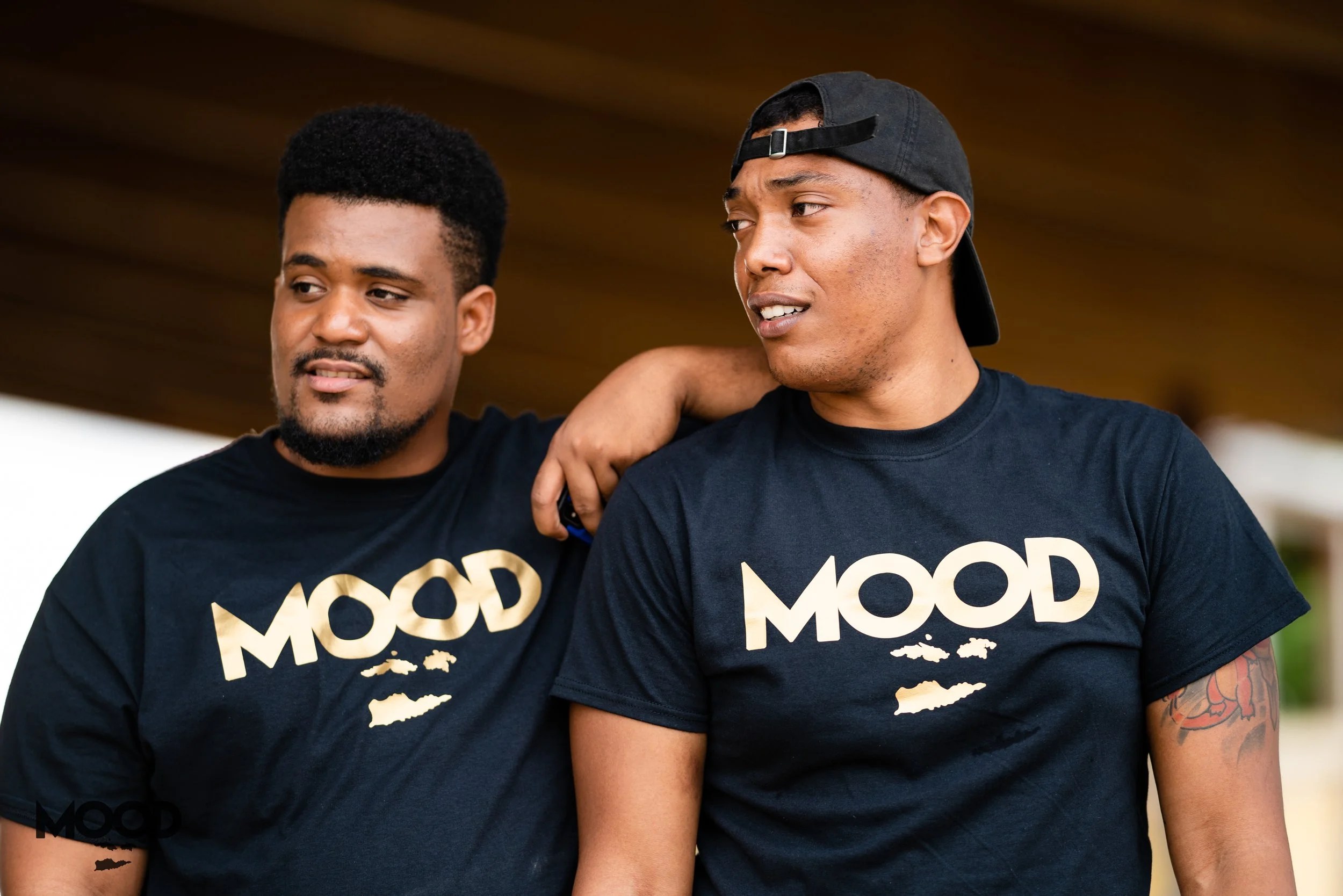 Two young men standing close together, wearing matching black t-shirts with the word 'MOOD' in large beige letters, outdoors with a blurred background.