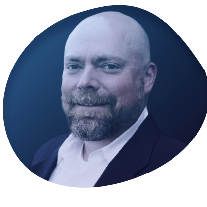 Portrait of a bald man with a beard wearing a suit and white shirt, standing in front of a brick wall.