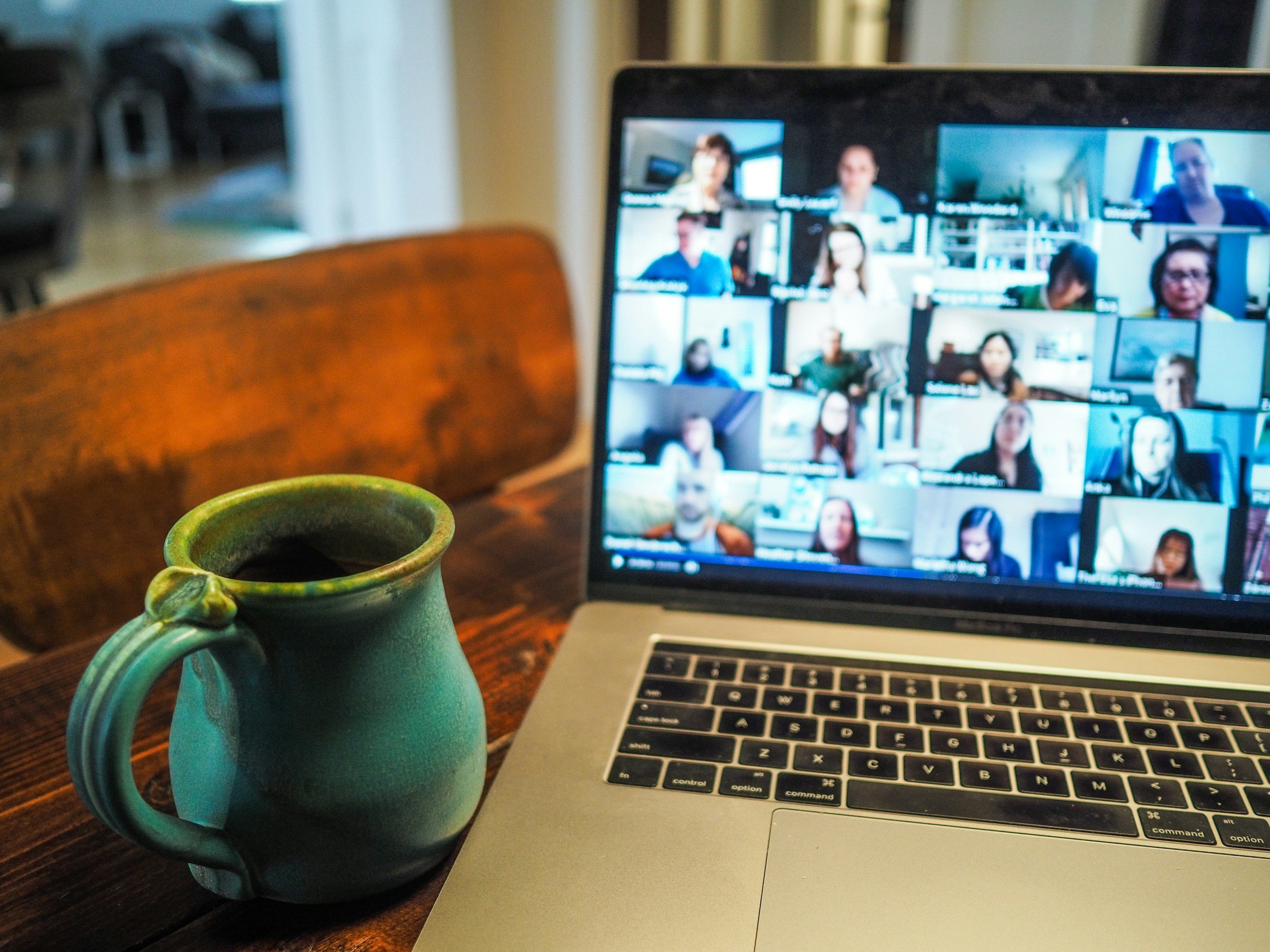 Zoomed-in view of a laptop screen displaying a video conference with multiple participants, placed on a wooden table next to a ceramic mug filled with coffee.