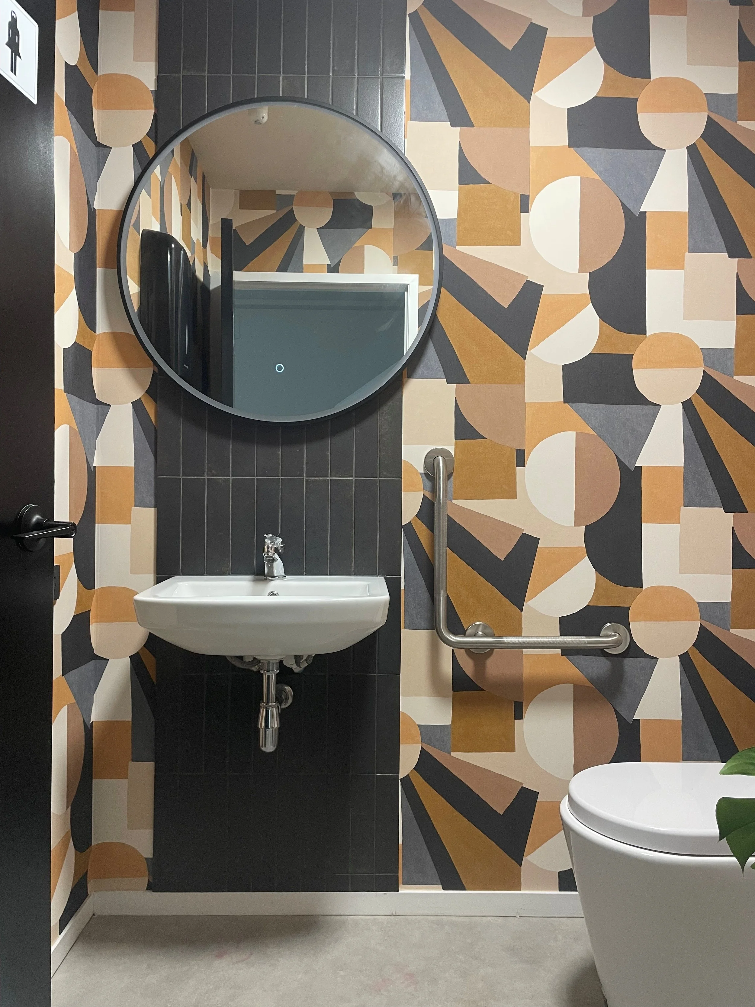 A modern bathroom with patterned geometric wallpaper, a circular mirror with LED lighting, a white sink, and a toilet. There is a black trash can on the floor and a person taking a photo in the mirror, with a man in a yellow shirt and hat reflected in the background.