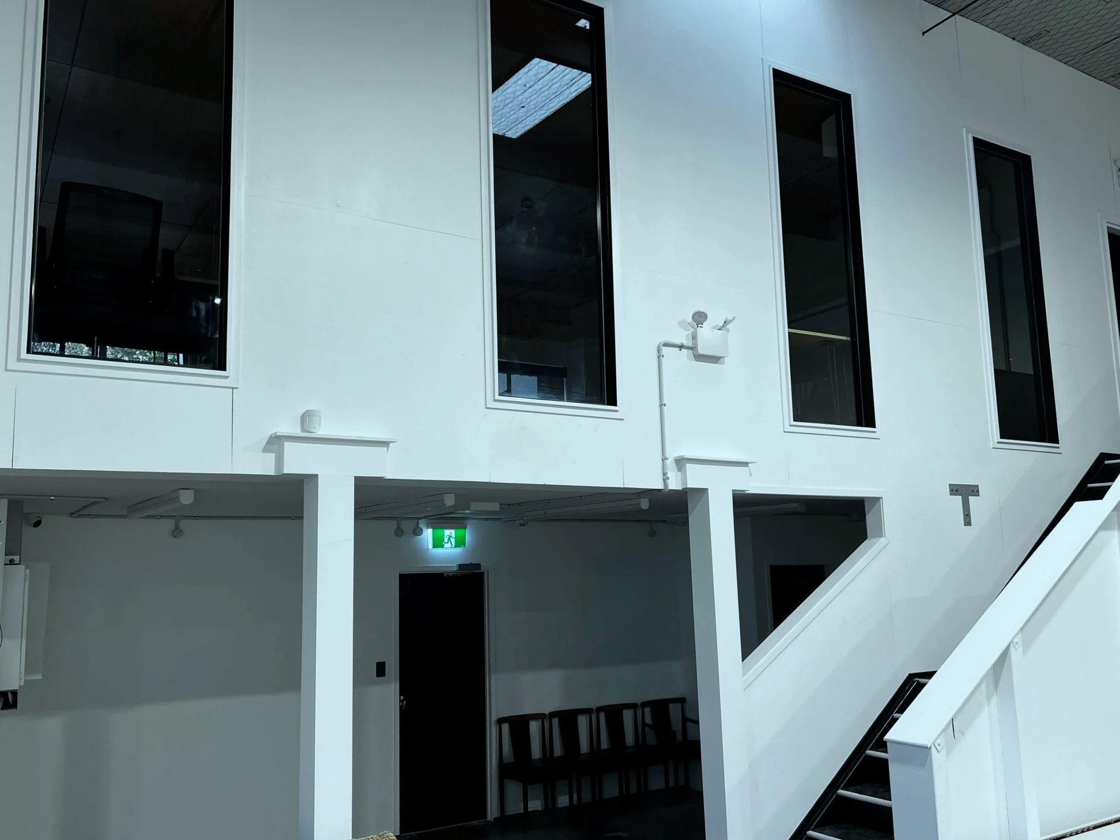 Interior of a modern building featuring high windows and a staircase with a black handrail. The walls are white, and there is an illuminated green emergency exit sign above a door.
