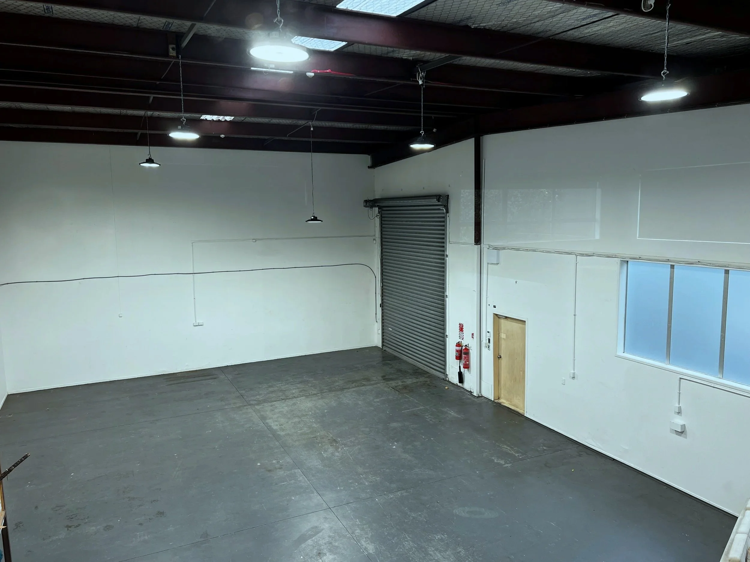 Interior of an empty warehouse with high ceiling, fluorescent lighting, a large metal roll-up door, a small wooden door, a window, and fire extinguishers.