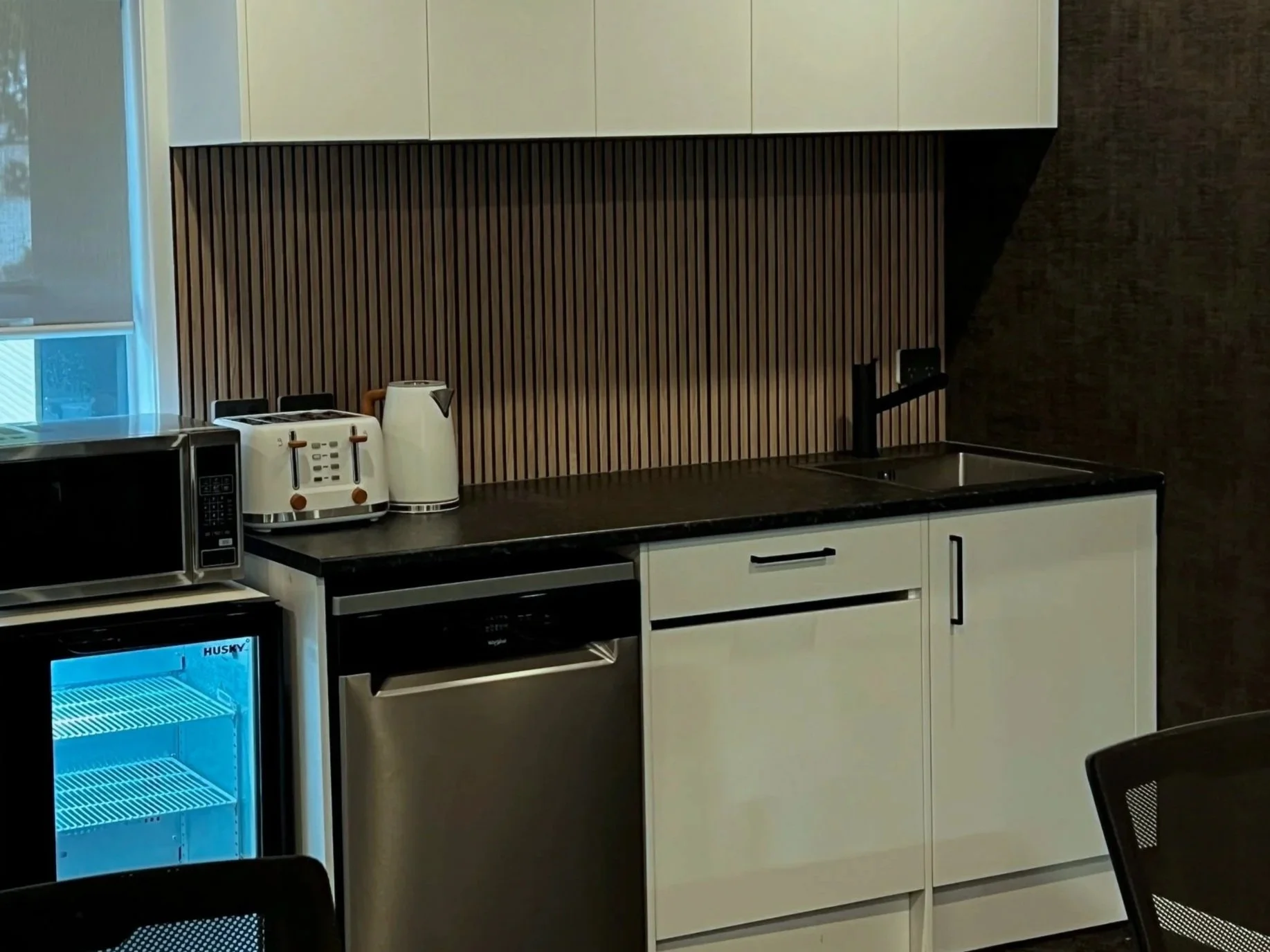 Modern kitchen with white cabinets, a black countertop, a stainless steel dishwasher, a mini fridge with a blue light, a microwave, a toaster, and a kettle. There is a window with blinds and a wooden slat backsplash.