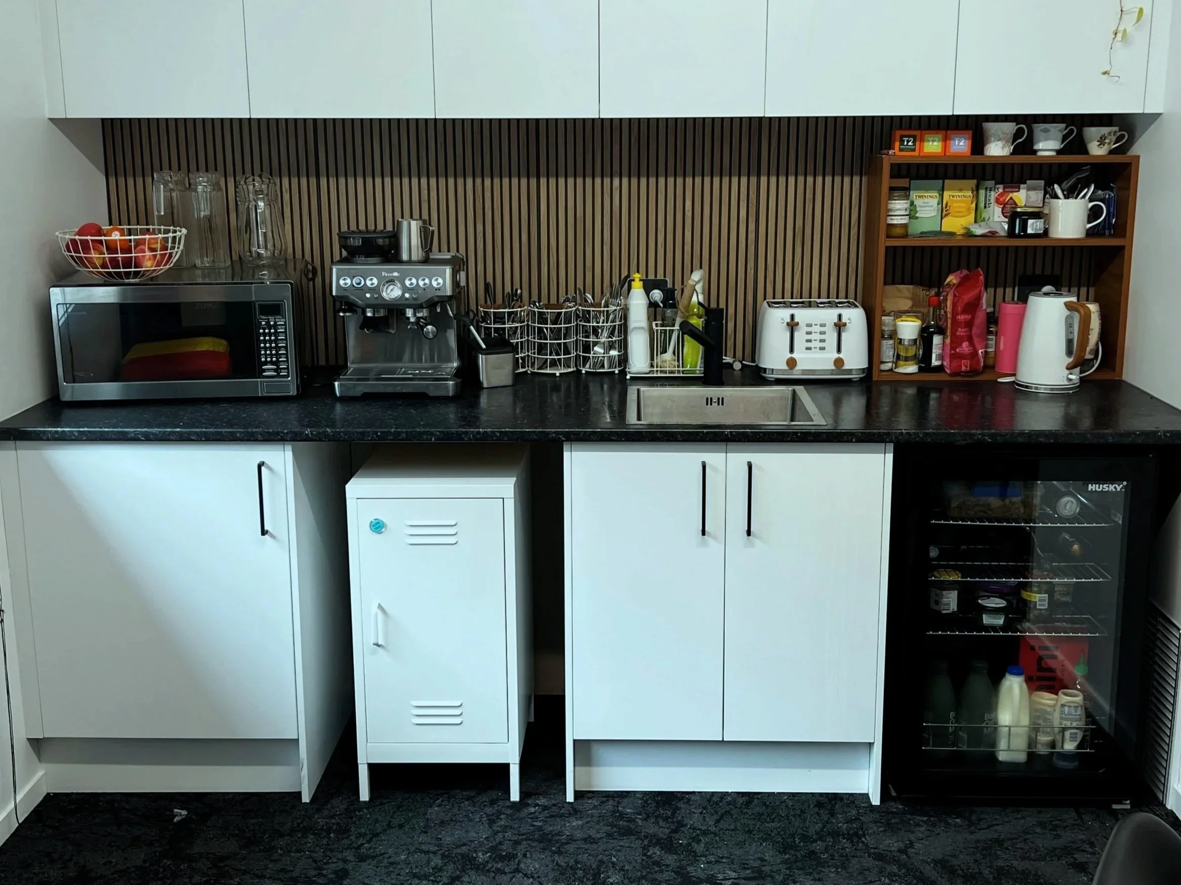 A modern kitchen counter with appliances including a microwave, espresso machine, toaster, and electric kettle, with a wooden shelf containing cups, tea, and snacks above. There is a sink with cleaning supplies, a mini fridge with beverages, and storage cabinets below.