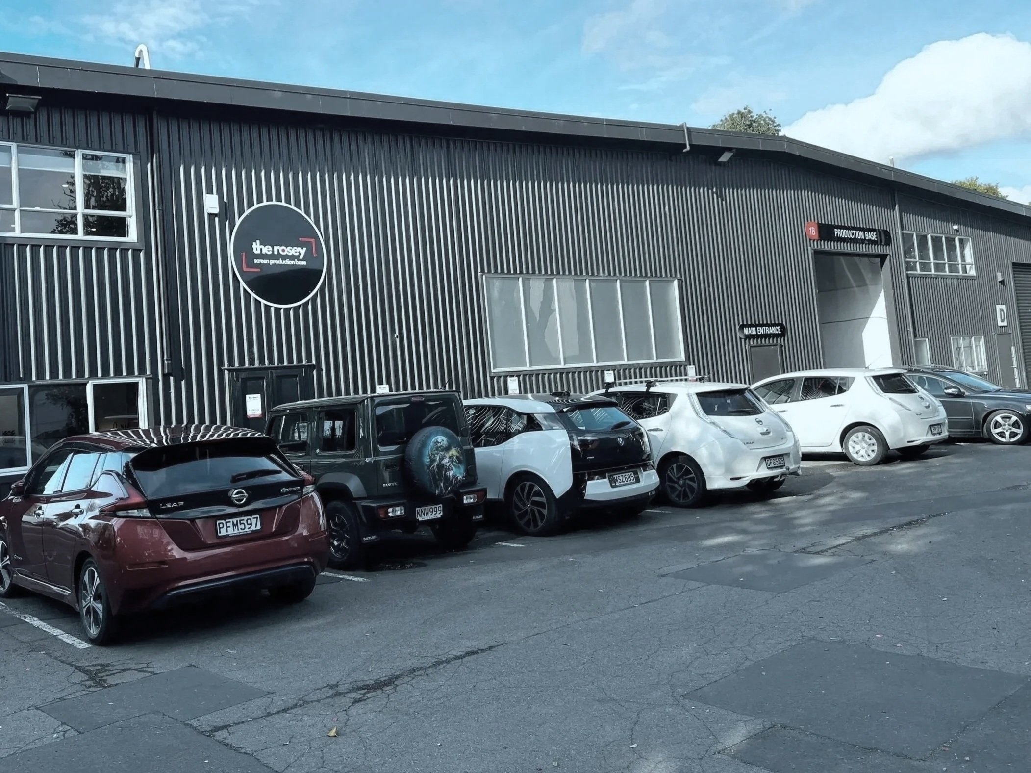 A parking lot in front of a black industrial-style building with multiple cars parked, blue sky with some clouds overhead, and trees in the background.