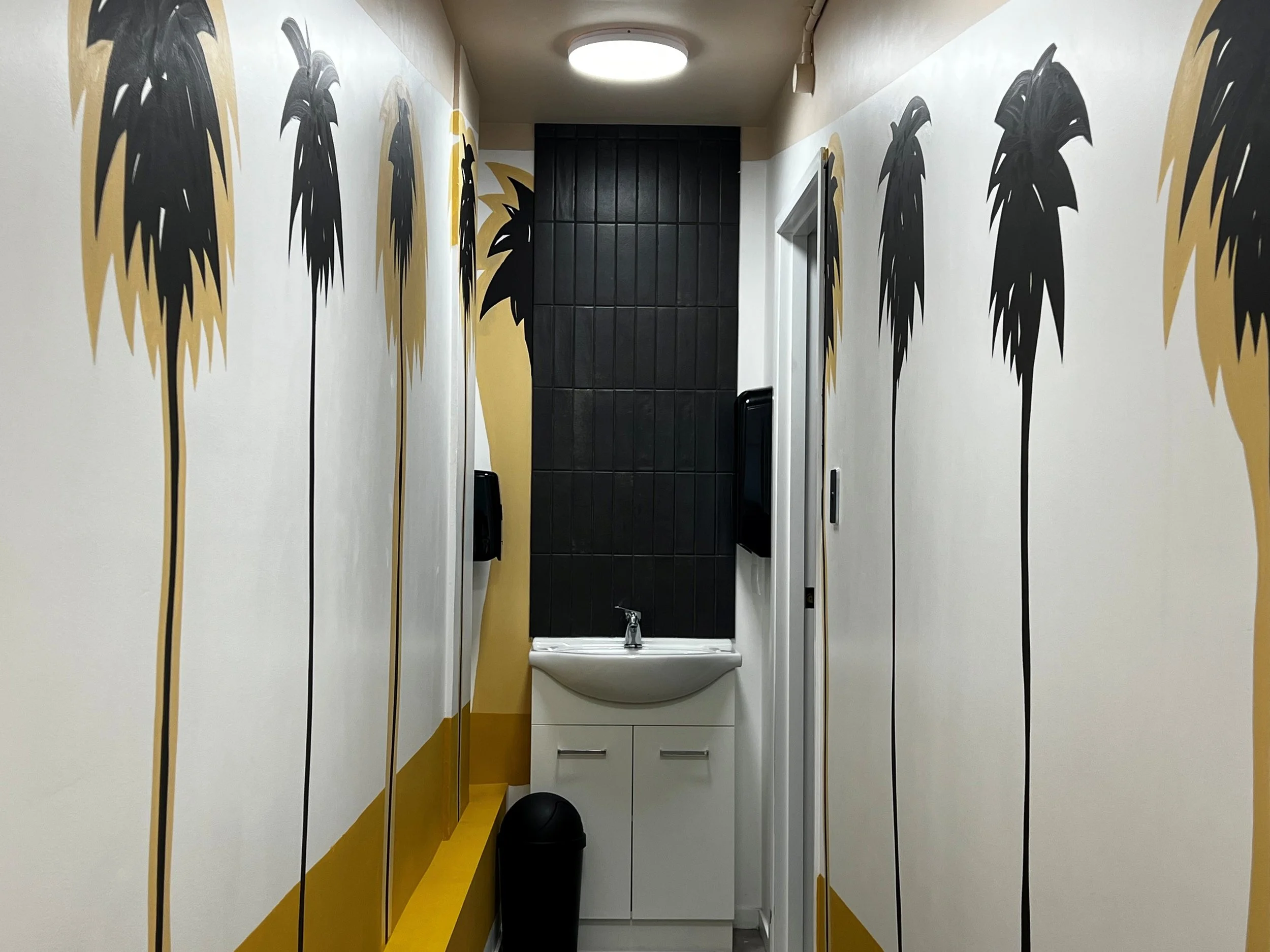 Narrow hallway with white walls decorated with large black and yellow palm trees, white sink with cabinets, black tile wall, black trash can, and wall-mounted paper towel dispenser.