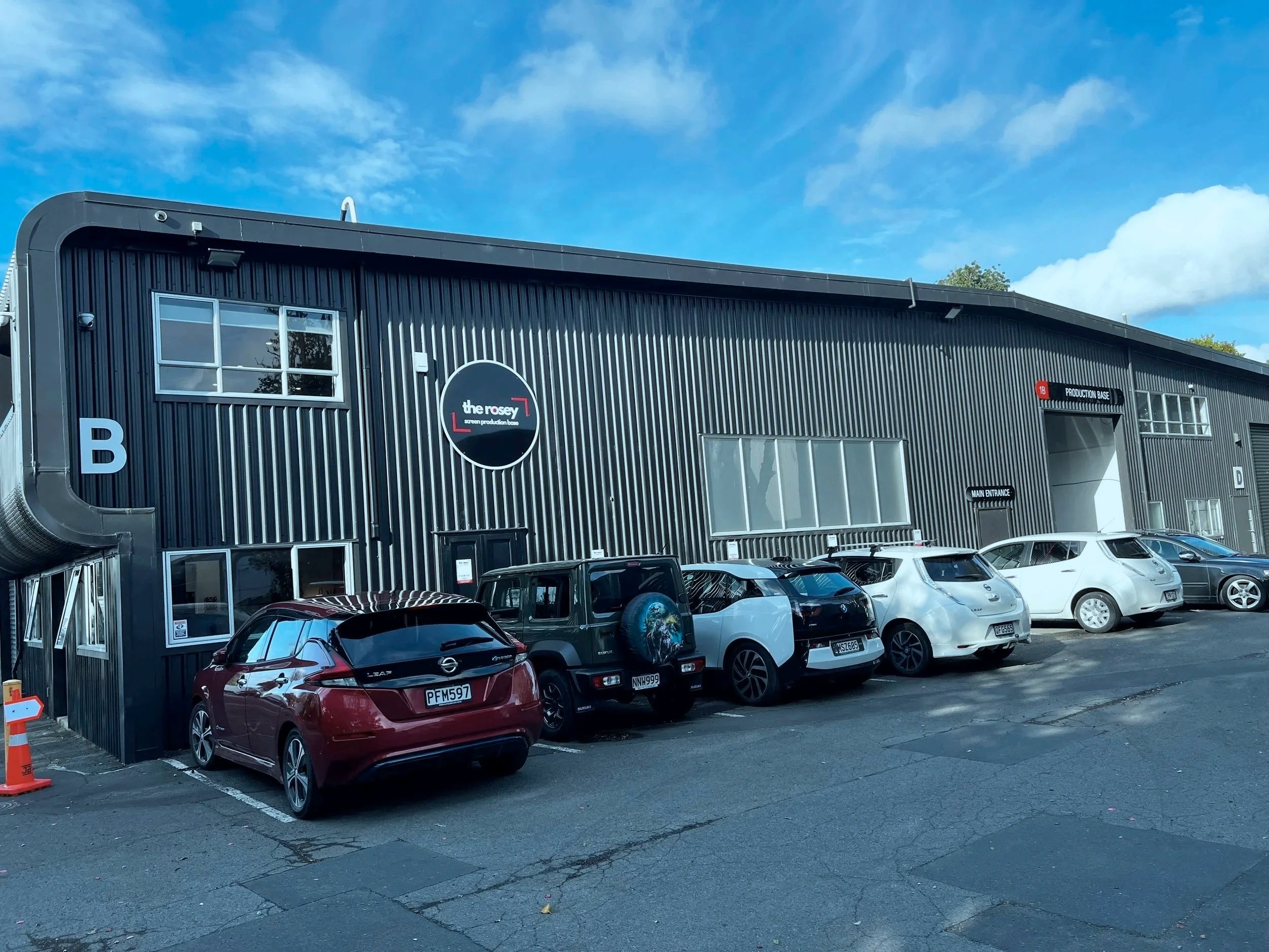 A parking lot in front of a black industrial-style building with multiple cars parked, blue sky with some clouds overhead, and trees in the background.