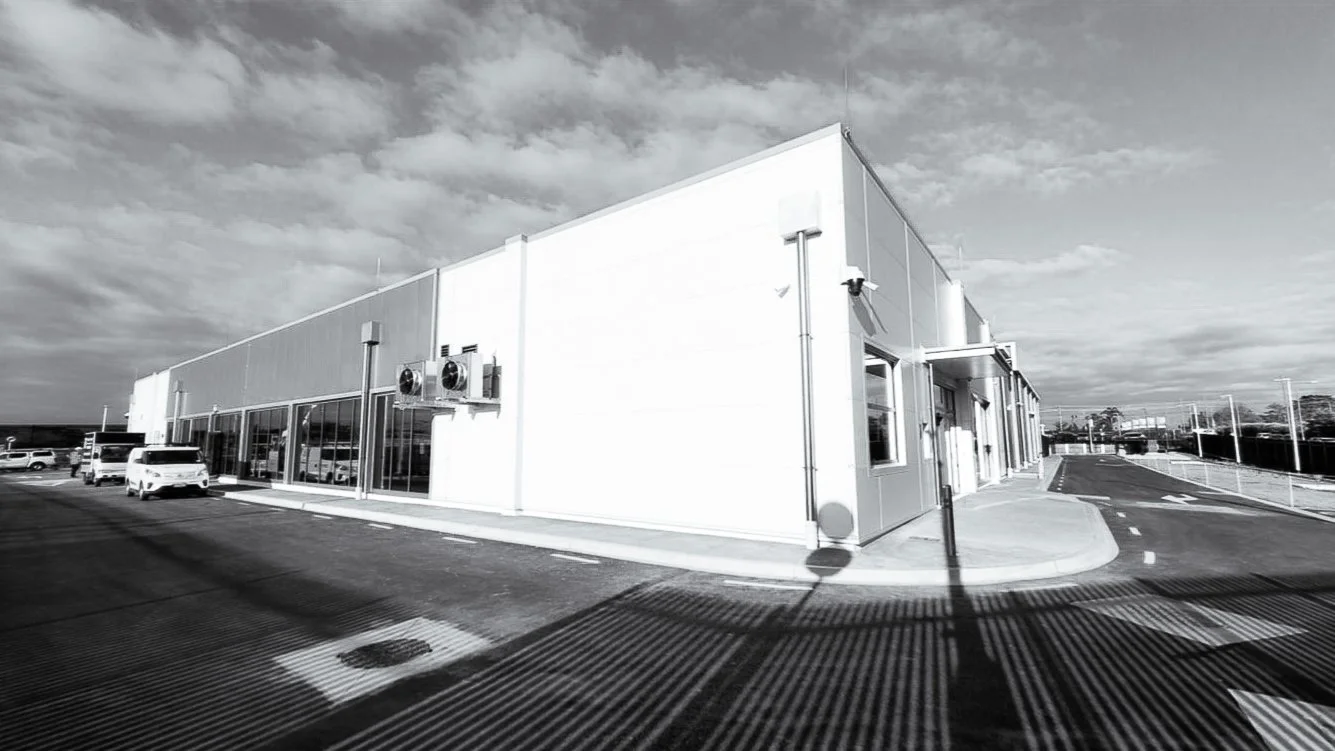 A modern commercial building with a large parking lot, several cars, and a cloudy sky in black and white.