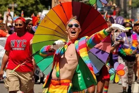 Person dressed in rainbow-colored costume holding a rainbow umbrella during a parade with a crowd of people in the background.