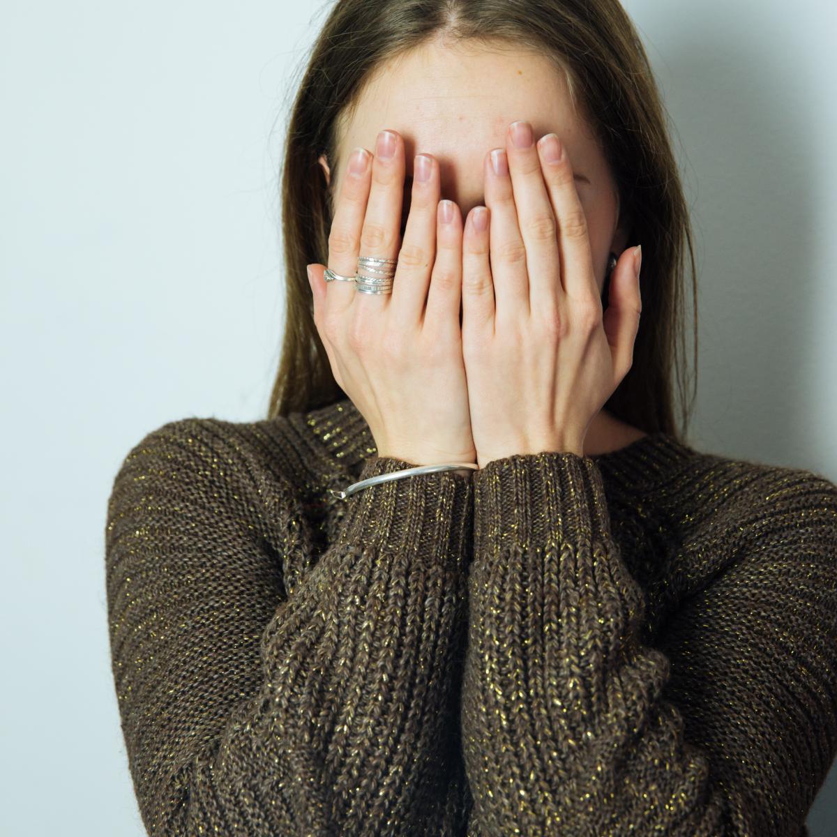 A young woman with brown hair hiding her face behind both hands, wearing a sparkly sweater, illustrating the emotional exhaustion of high-masking ADHD in women.