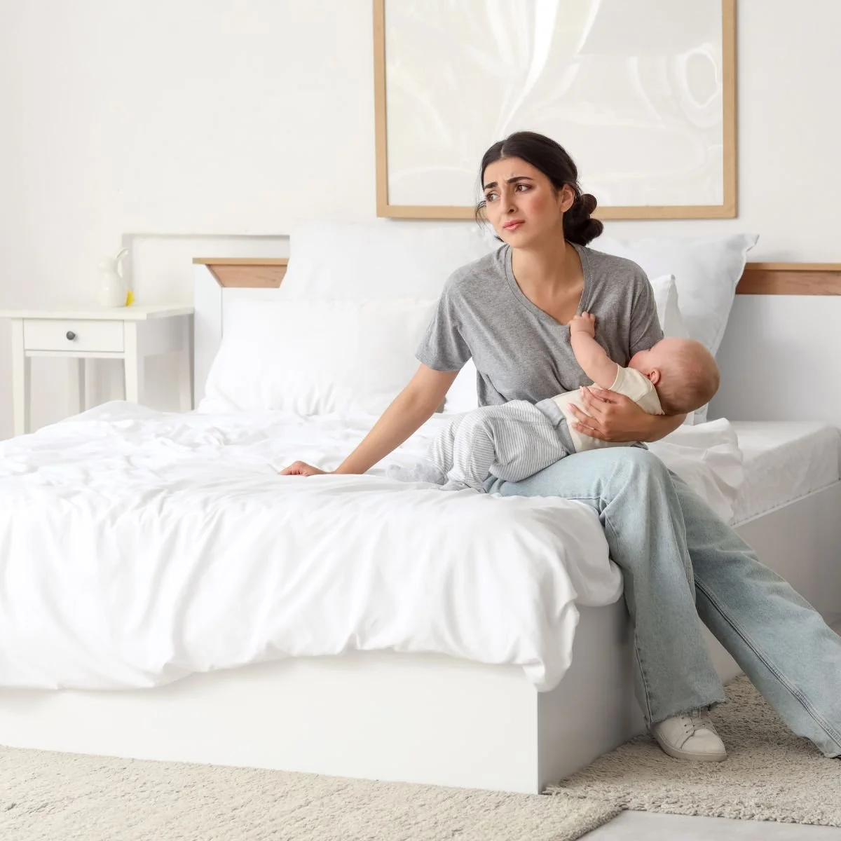 Exhausted mother holding sleeping newborn on couch surrounded by baby care items during postpartum period