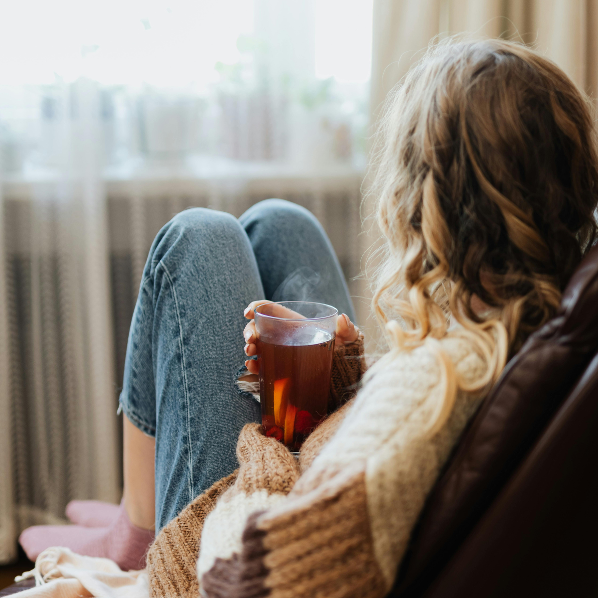 Rear view of a woman with curly blonde hair relaxing in an armchair and holding a hot mug of tea, representing the peace and relief that can come from a late ADHD diagnosis.