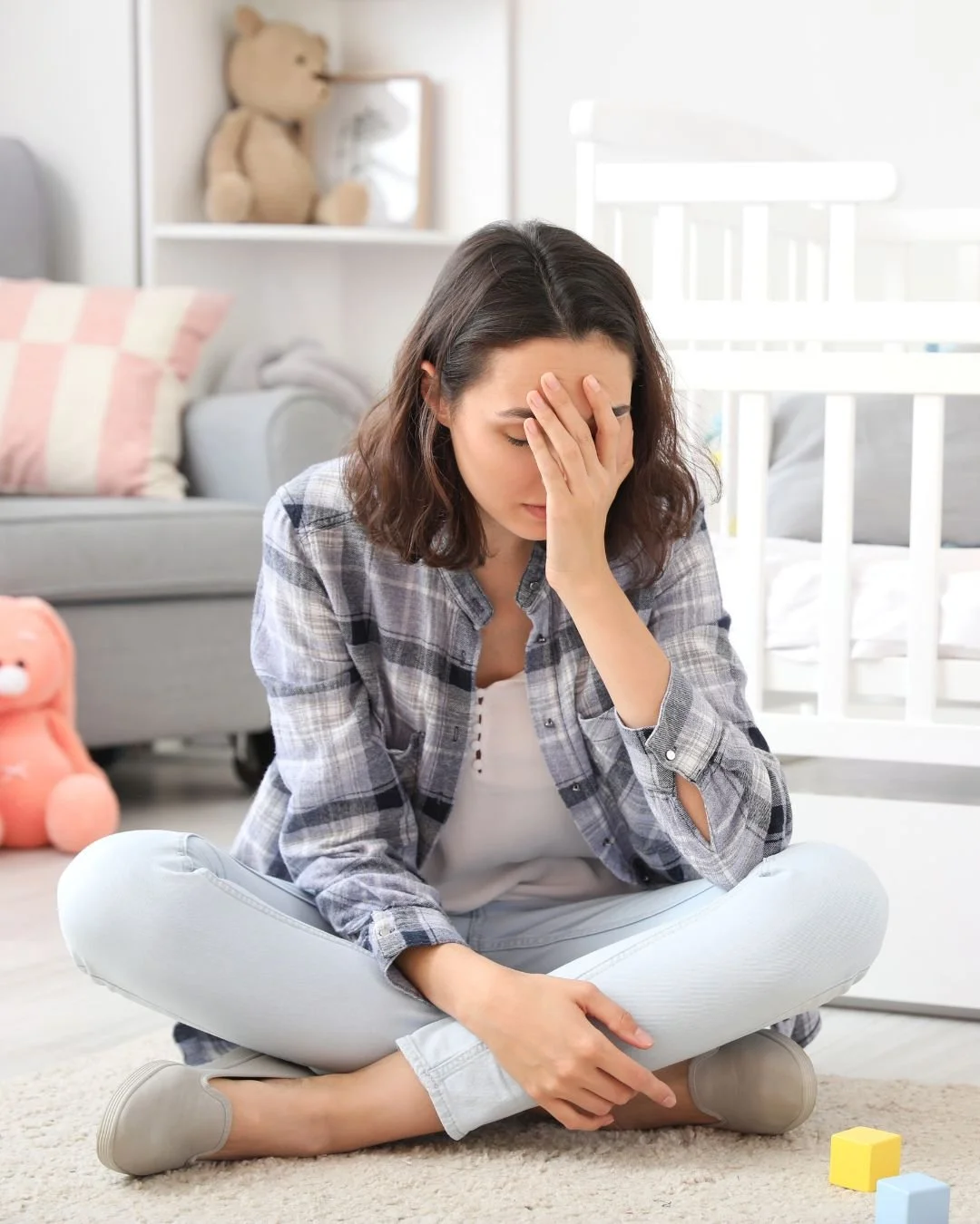 A woman sitting cross-legged on a carpet in a living room, holding her forehead with one hand and looking down, appearing stressed or upset.