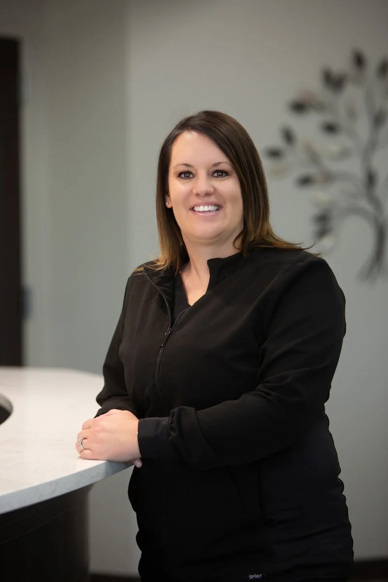 A woman with shoulder-length dark hair, smiling, wearing a black top against a white background.