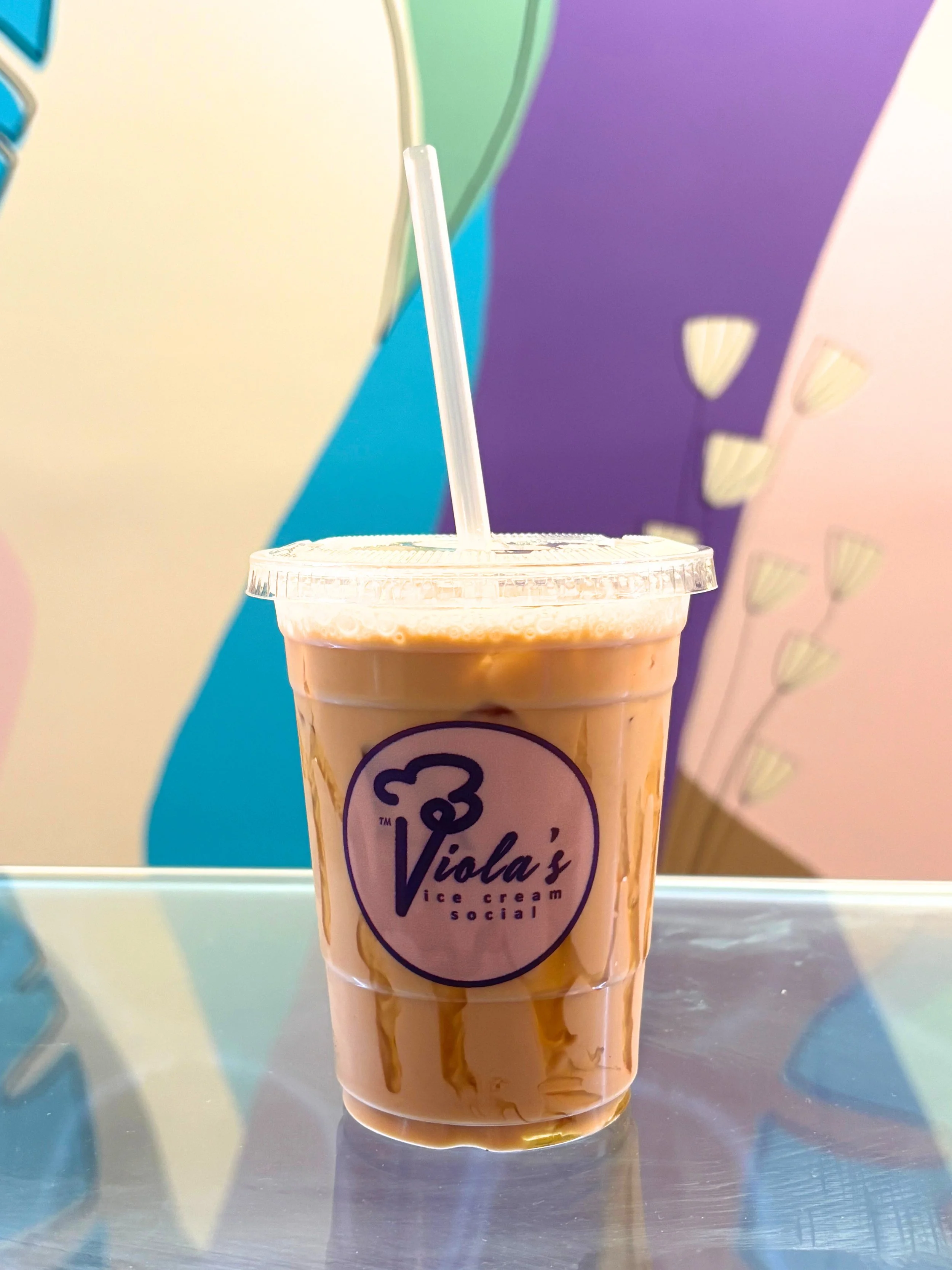 A plastic cup of Viola's ice cream social with caramel sauce drizzled on top, placed on a reflective surface with a colorful abstract background.