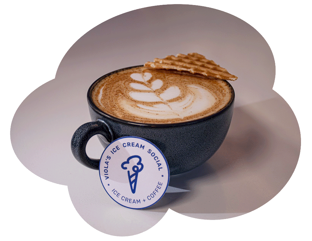 A black mug of coffee with latte art and a waffle on the rim, next to a Viola's Ice Cream Social sticker.