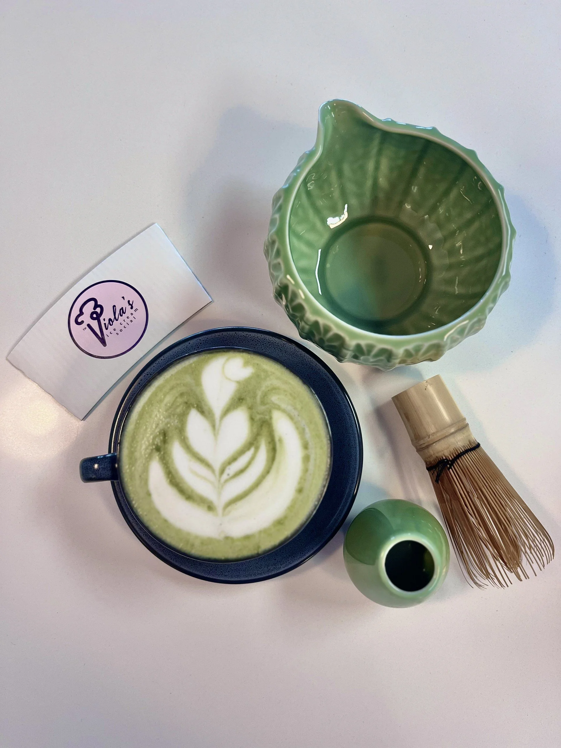 A cup of matcha latte with latte art, a green ceramic pitcher, a small green bowl, a bamboo whisk, and a white card with a purple logo on a white surface.