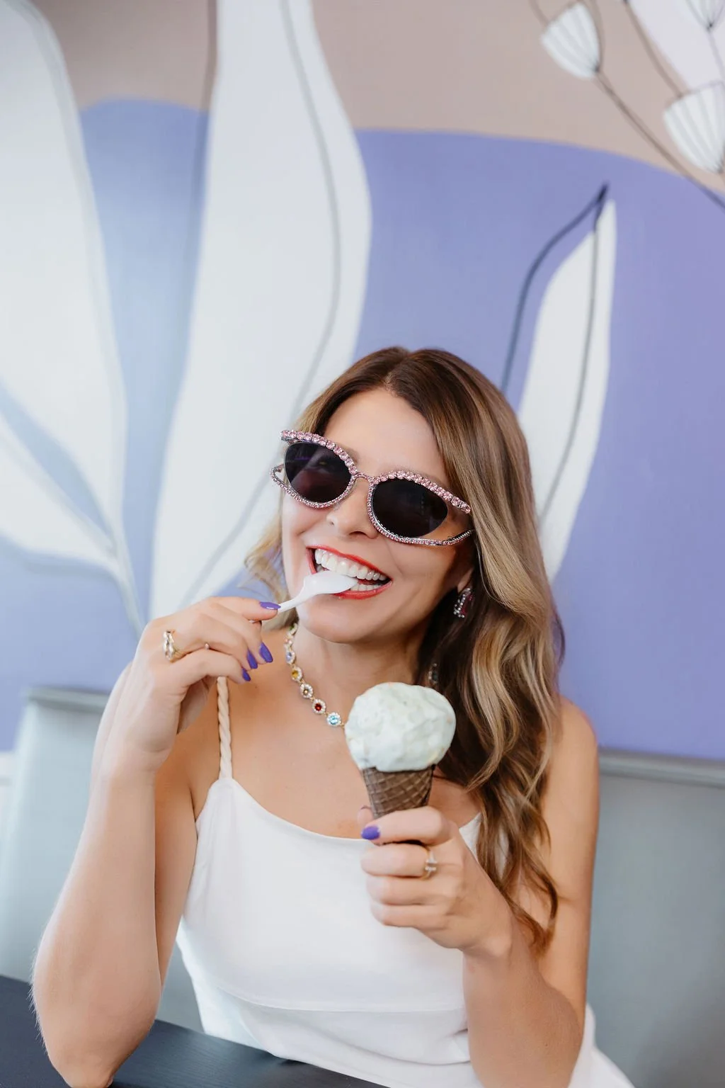 A woman with wavy, light brown hair wearing large, dark sunglasses with rhinestone embellishments, enjoying an ice cream cone while smiling at the camera. She is dressed in a white top, accessorized with a colorful necklace, earrings, and a ring, with purple nail polish, in a decorated indoor setting.