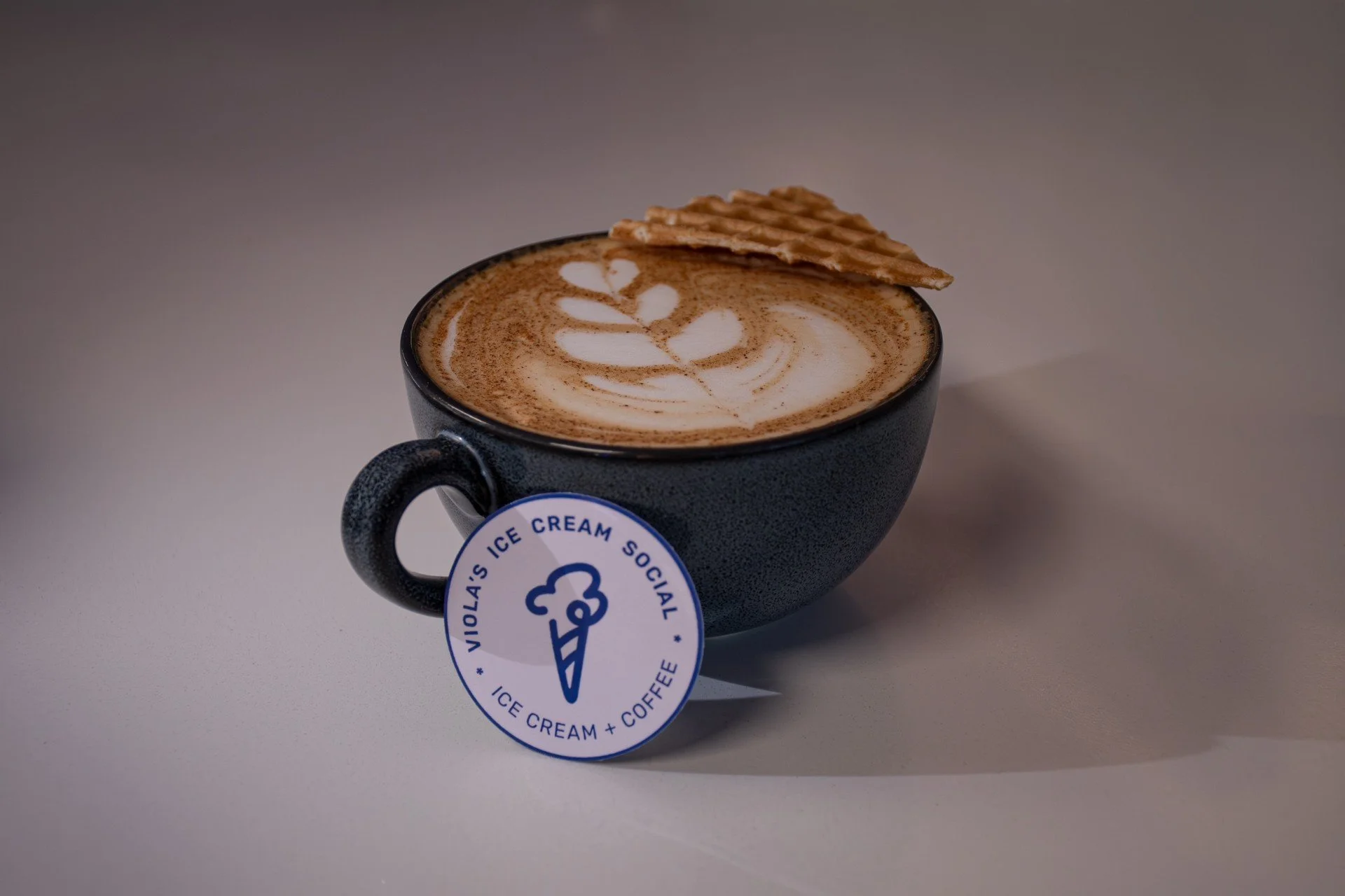 A black mug filled with coffee topped with latte art and a waffle cookie on the rim. The mug has a blue and white sticker saying 'Nolia's Ice Cream Social' with an ice cream cone logo.