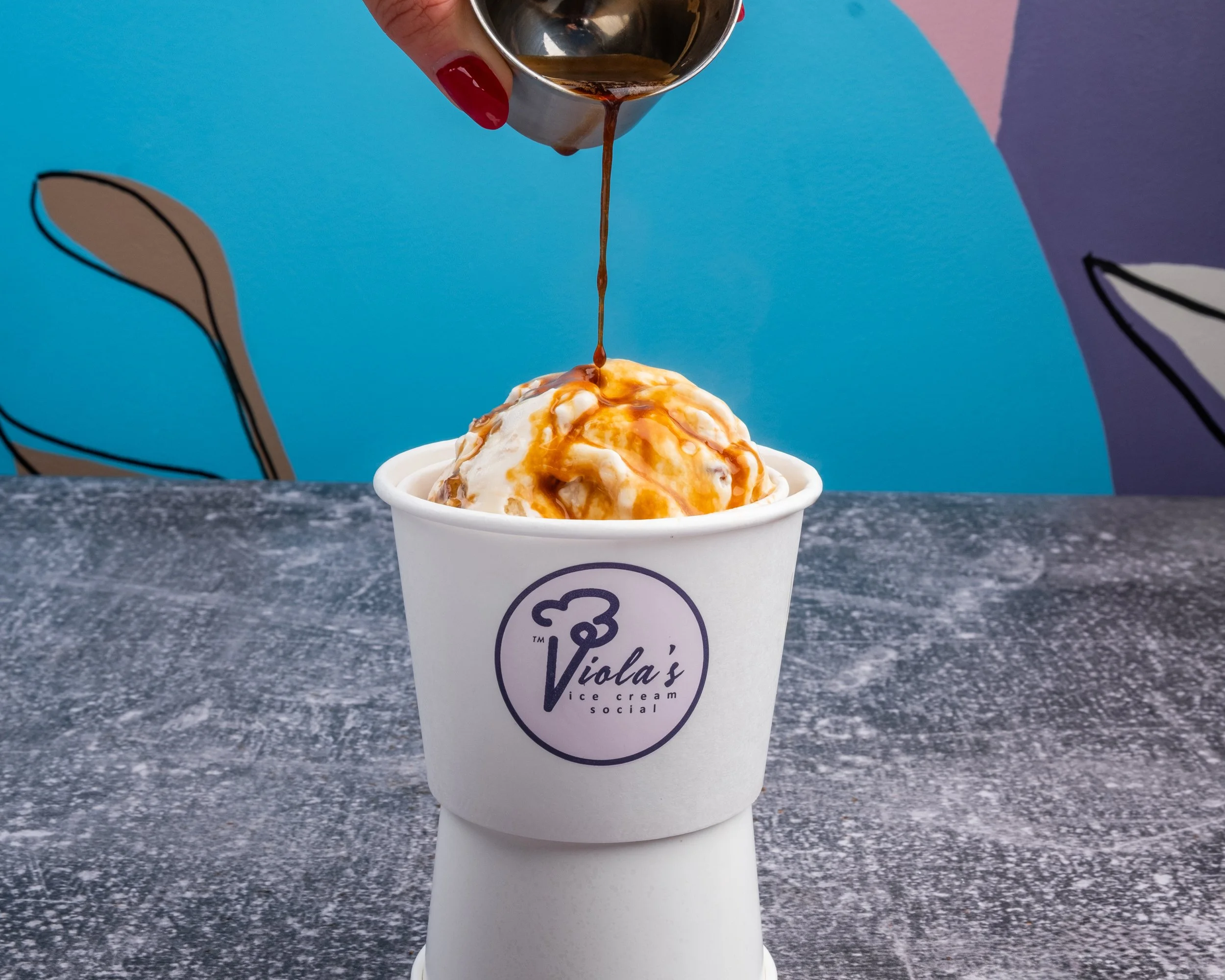 A cup of ice cream with caramel sauce, with hot fudge being poured over from a metal container held by a person with red nail polish. The cup has a logo that reads "Viola's ice cream social". The background includes a colorful, abstract wall with shades of blue, pink, and purple.