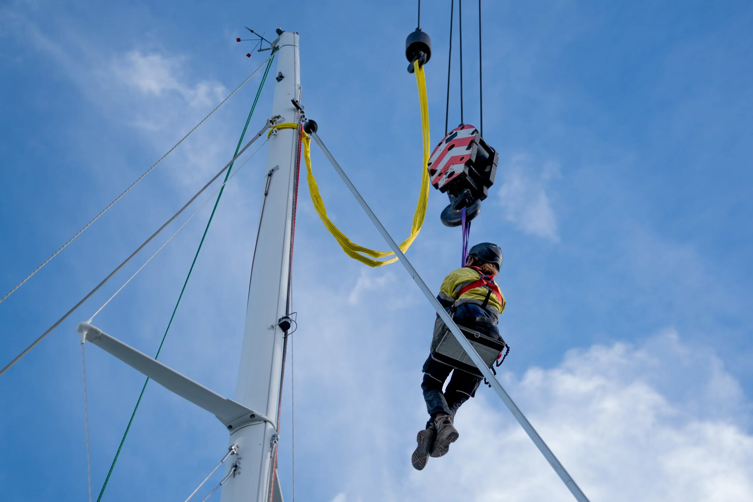 A person wearing a safety harness and helmet working on a tall sailboat mast against a blue sky with clouds, using a lift to reach the upper parts of the mast.