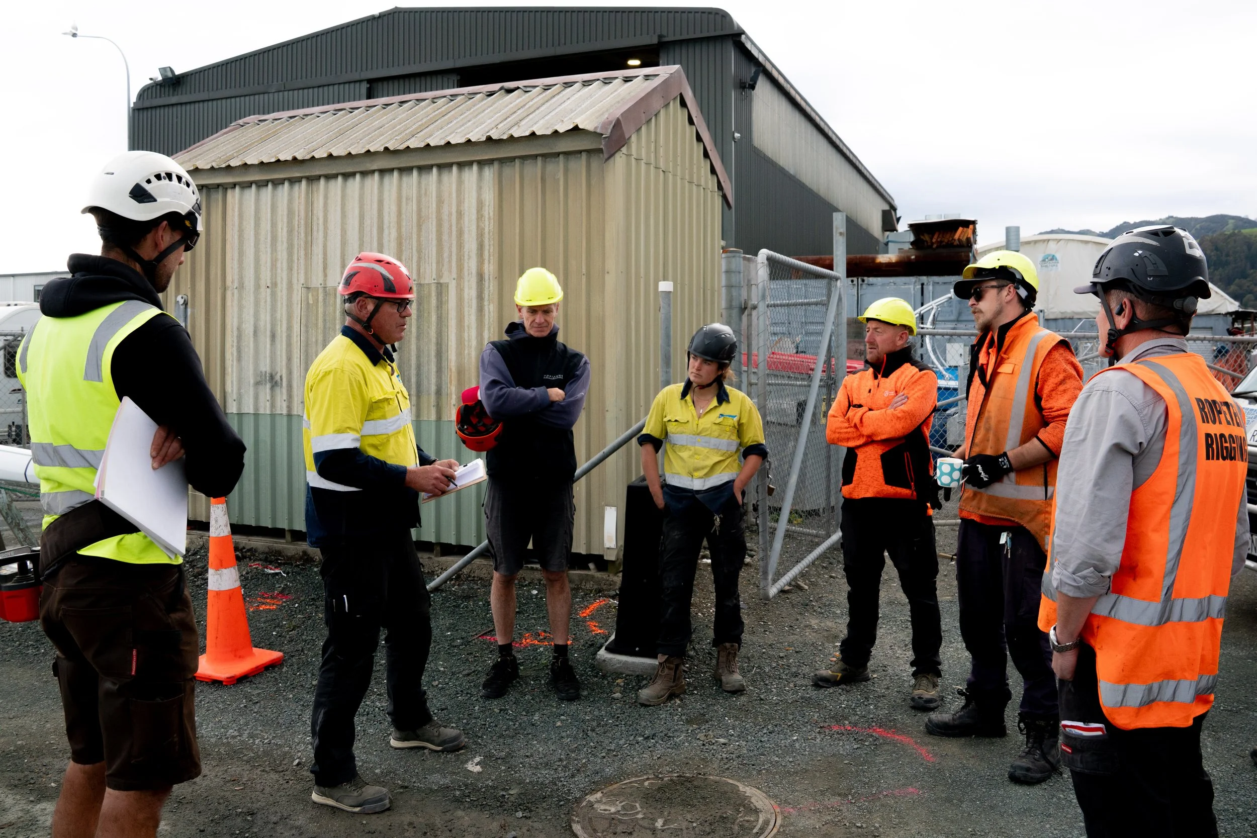 Group of construction workers and engineers gathered outdoors near industrial buildings, wearing safety gear including helmets and vests, engaged in a discussion or briefing.