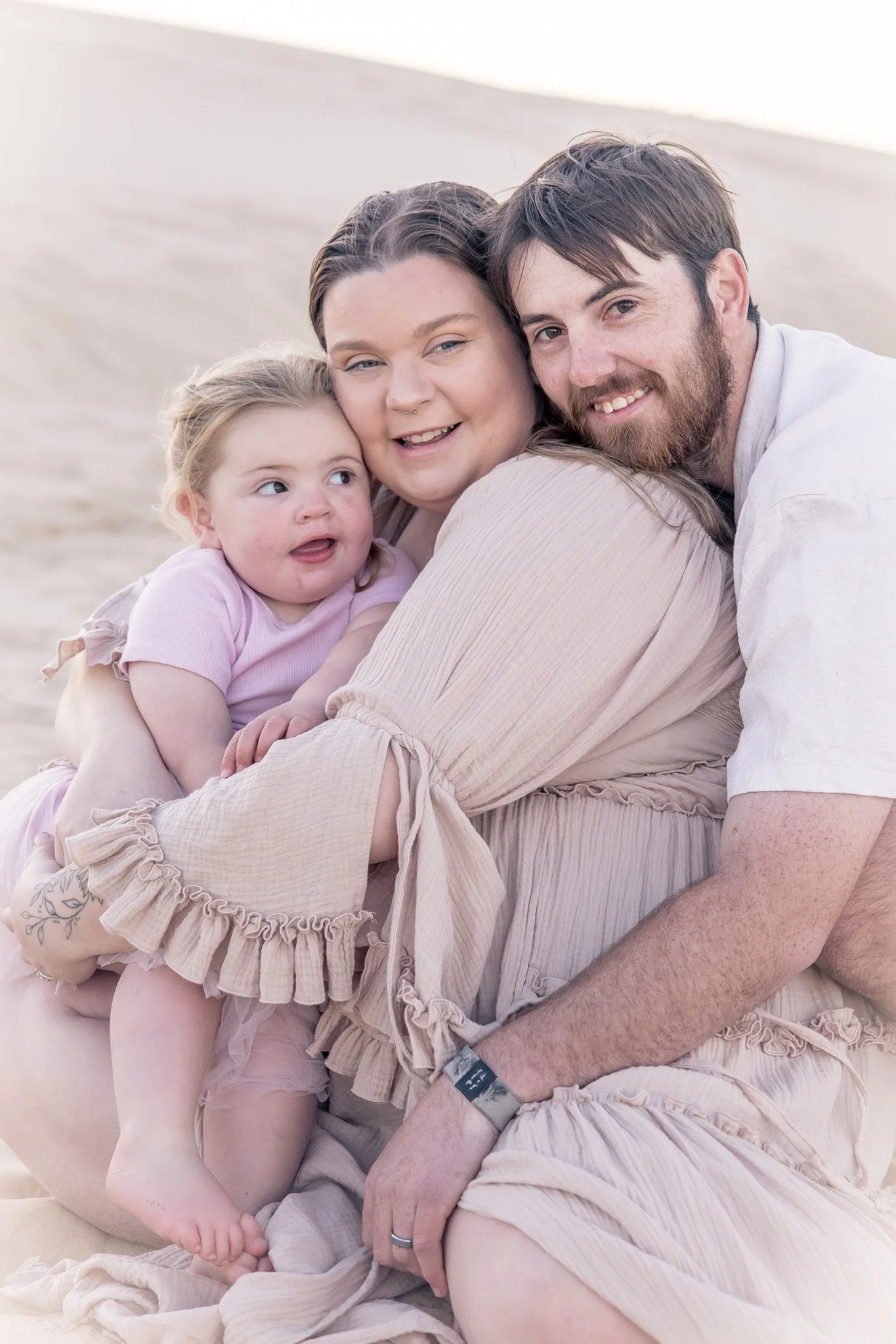 Family of three sitting on a sandy beach, smiling and cuddling, with a blurred background of sand and sky.