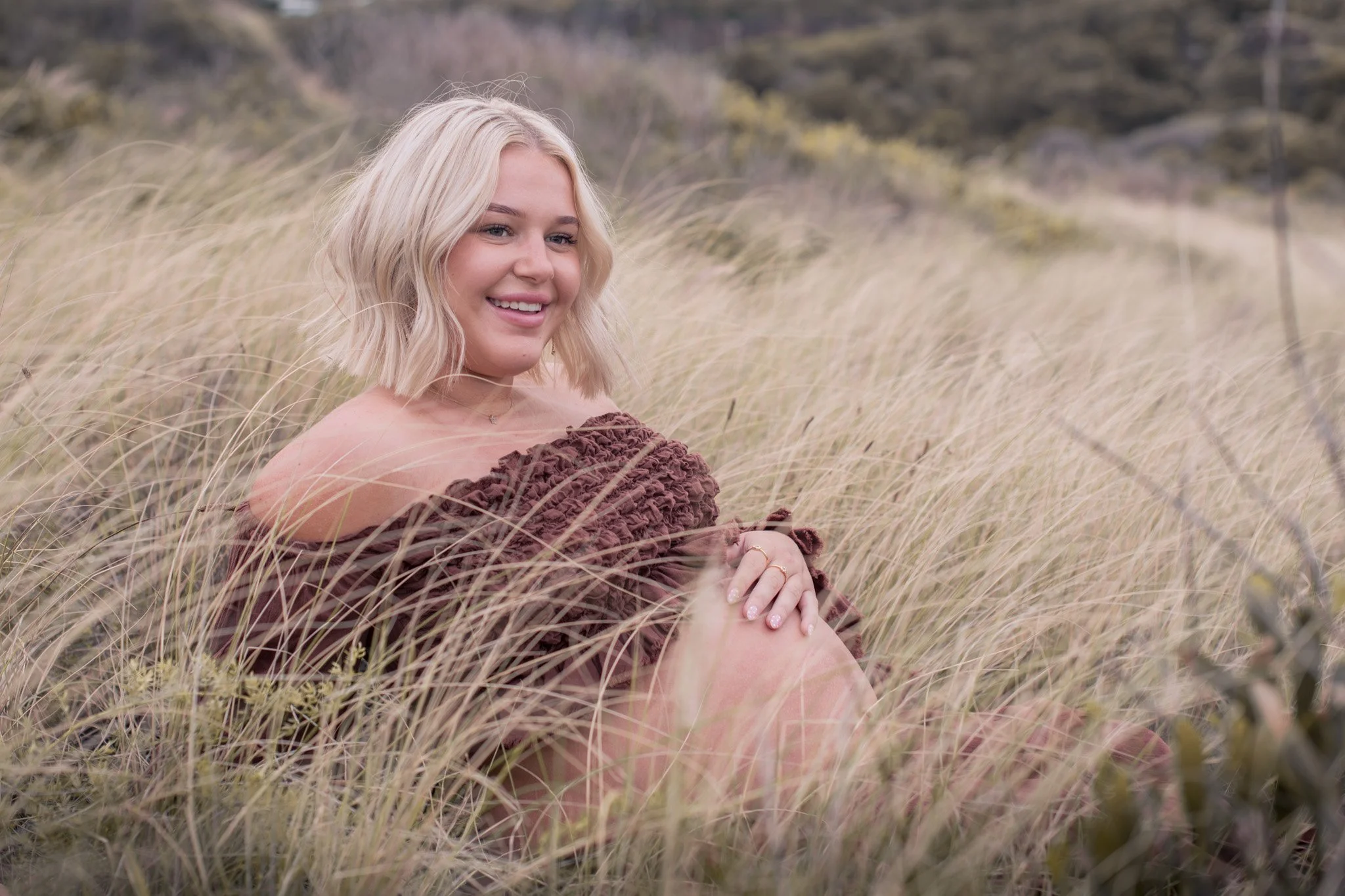 A young woman with blonde hair and a brown off-the-shoulder dress sitting in tall grass in an outdoor setting with trees and foliage in the background.