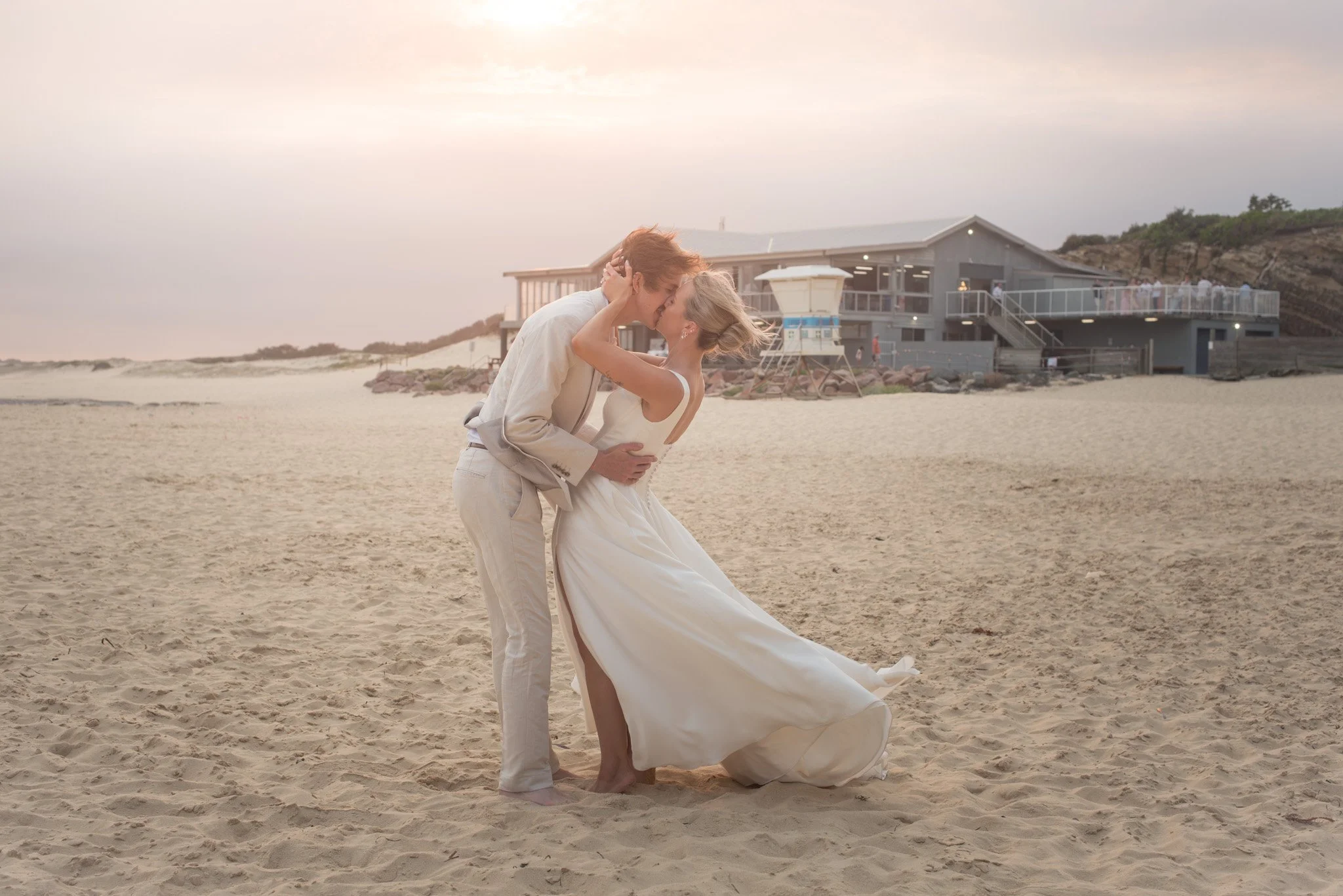 A couple dressed in wedding attire embracing and kissing on a sandy beach during sunset with a beach house in the background.