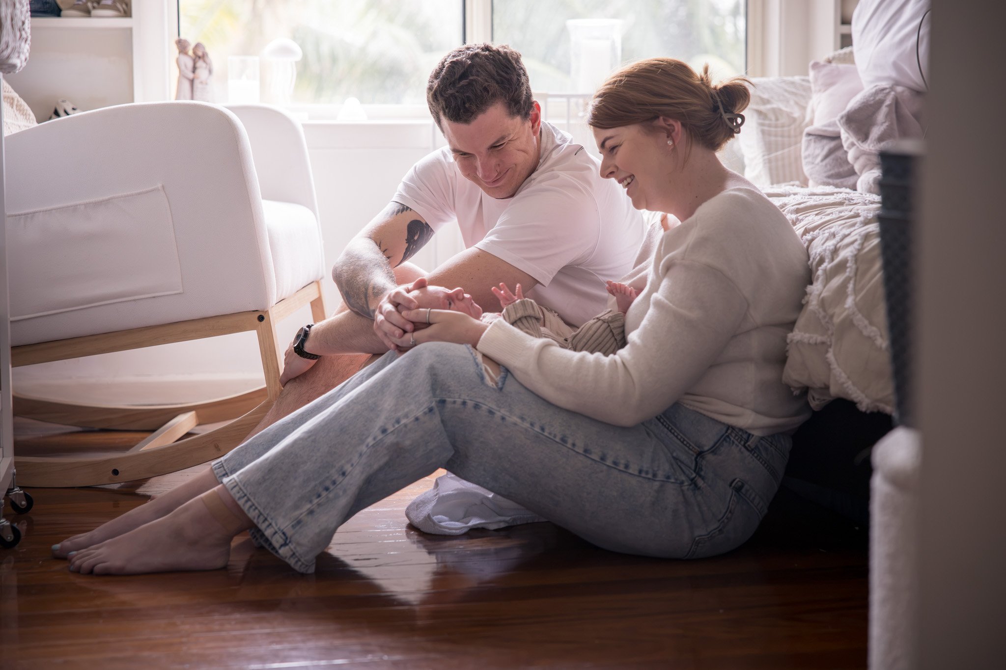 A couple sitting on the hardwood floor next to a bed, looking down at a baby who is lying on their side, with the woman holding the baby's hand. The room has natural light from a window behind them.