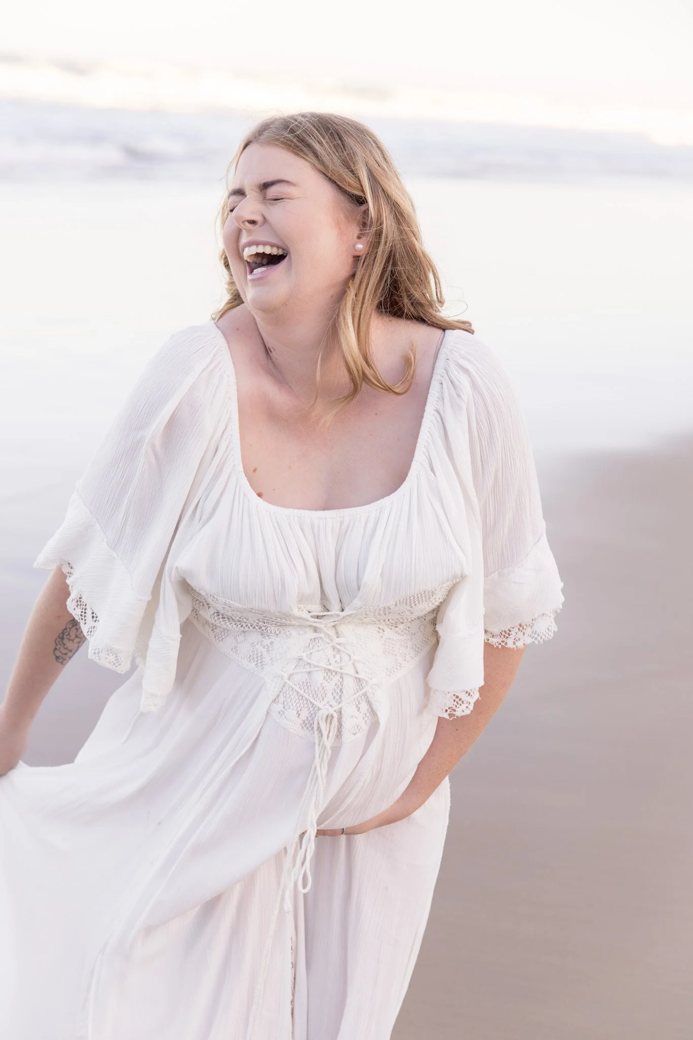A woman with red hair laughing on the beach, wearing a white dress with lace and embroidery details.