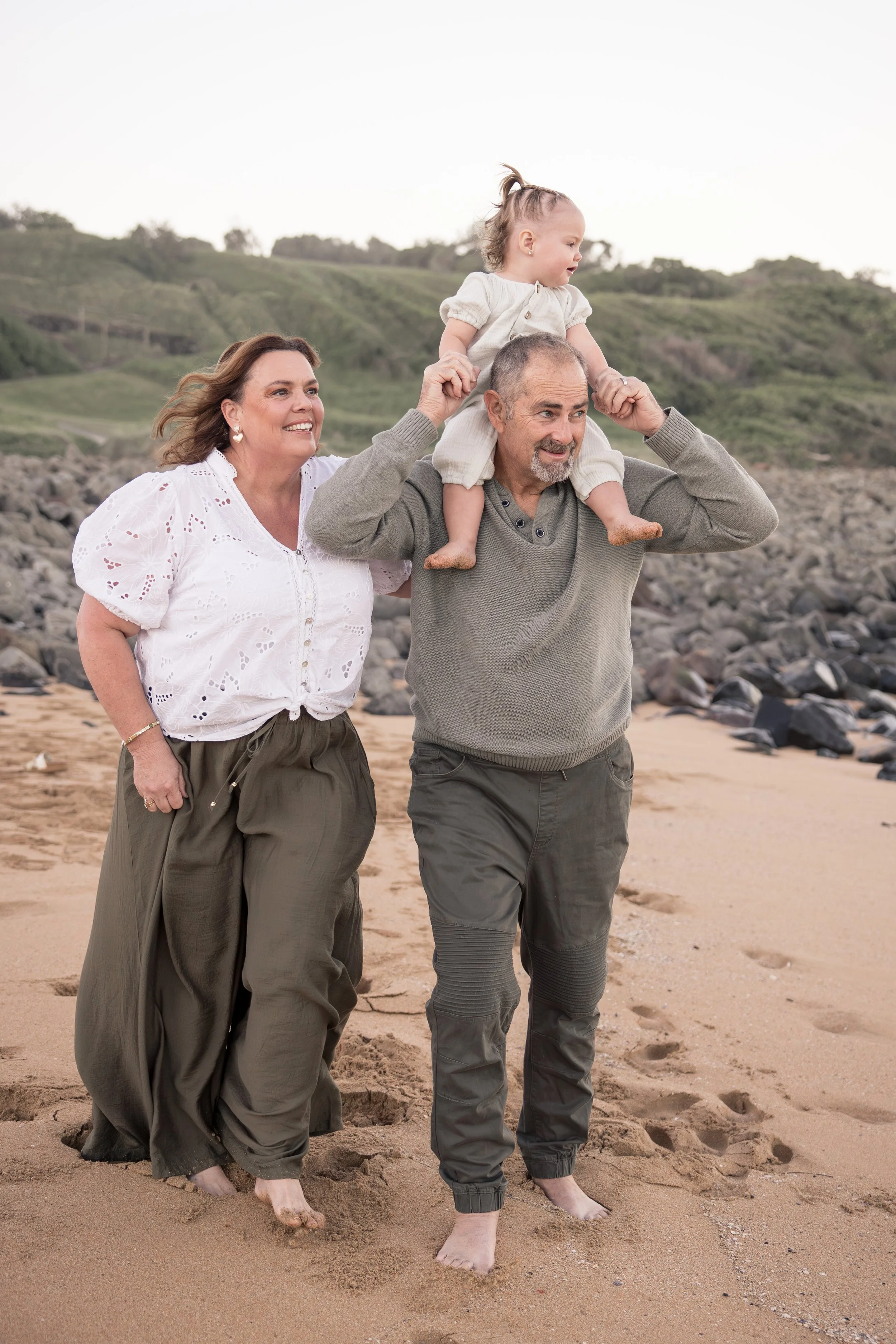 A family of three walking on the beach with the father carrying a young girl on his shoulders. The mother is walking beside them, all smiling and enjoying the outdoor scene with hills and rocky shoreline in the background.