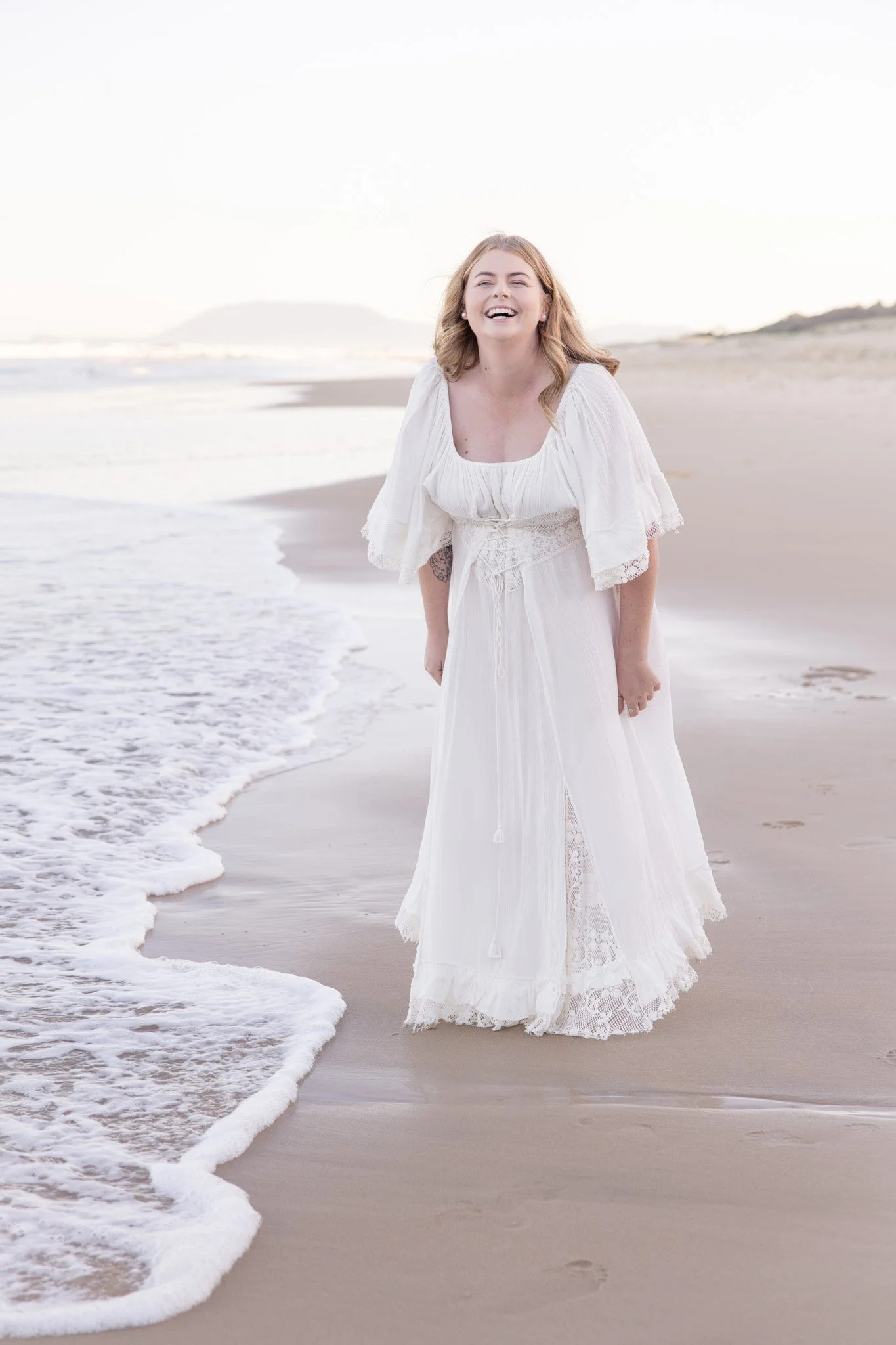 A woman in a white dress standing on a sandy beach with ocean waves and a distant landmass in the background, smiling and laughing.