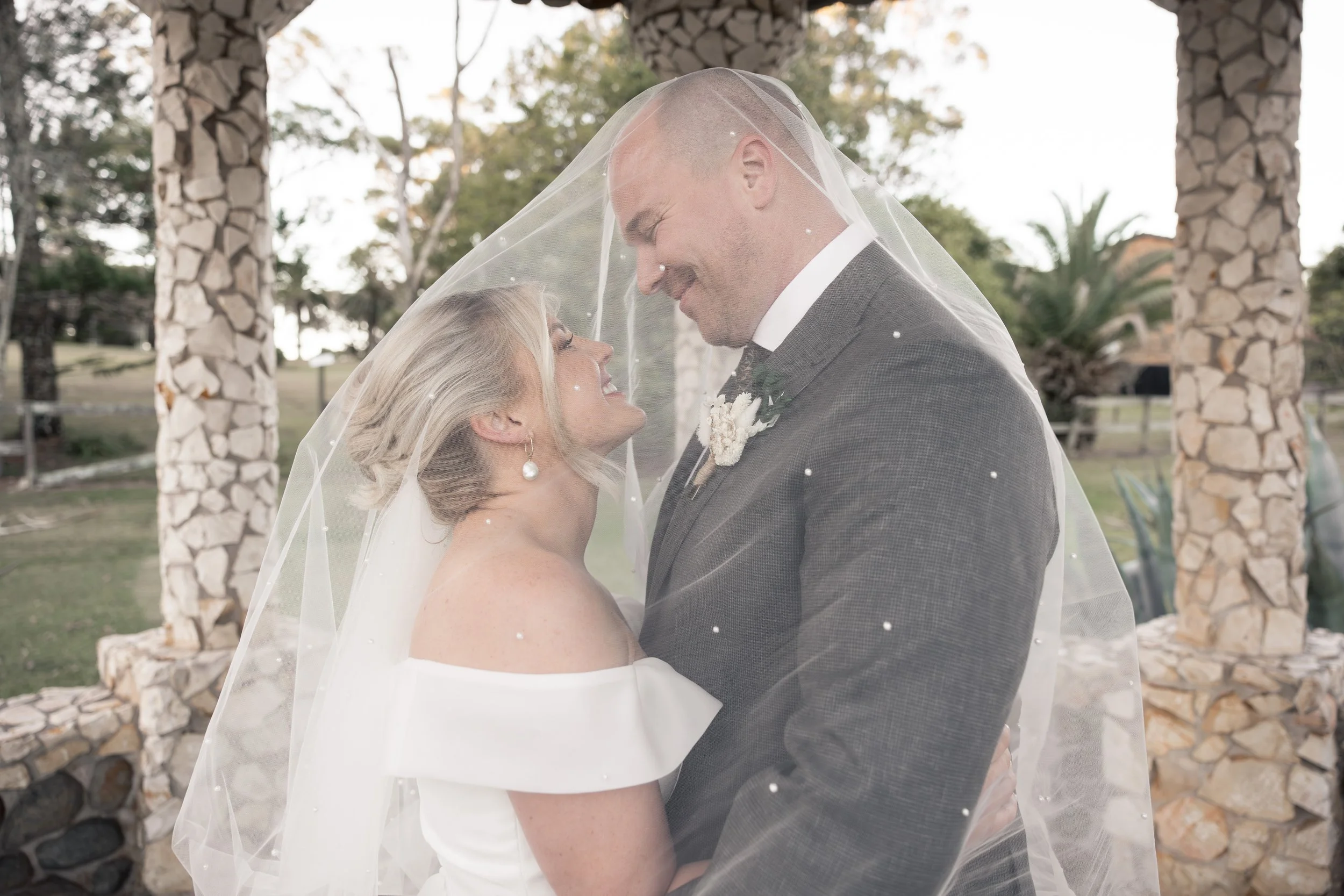 A bride and groom are standing close under a veil outdoors, smiling at each other, with stone pillars and trees in the background.