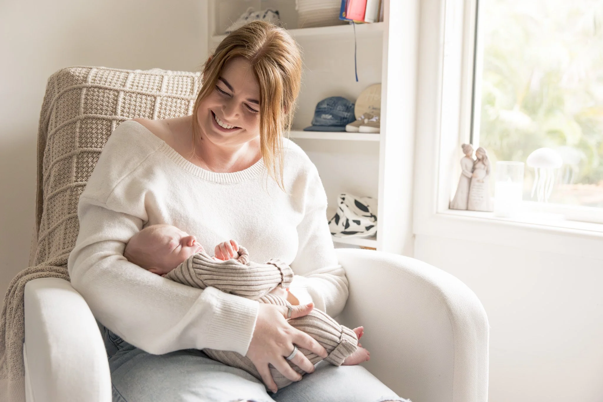 A woman holding a sleeping baby in a cozy room with sunlight through a window, smiling and looking down at the infant.