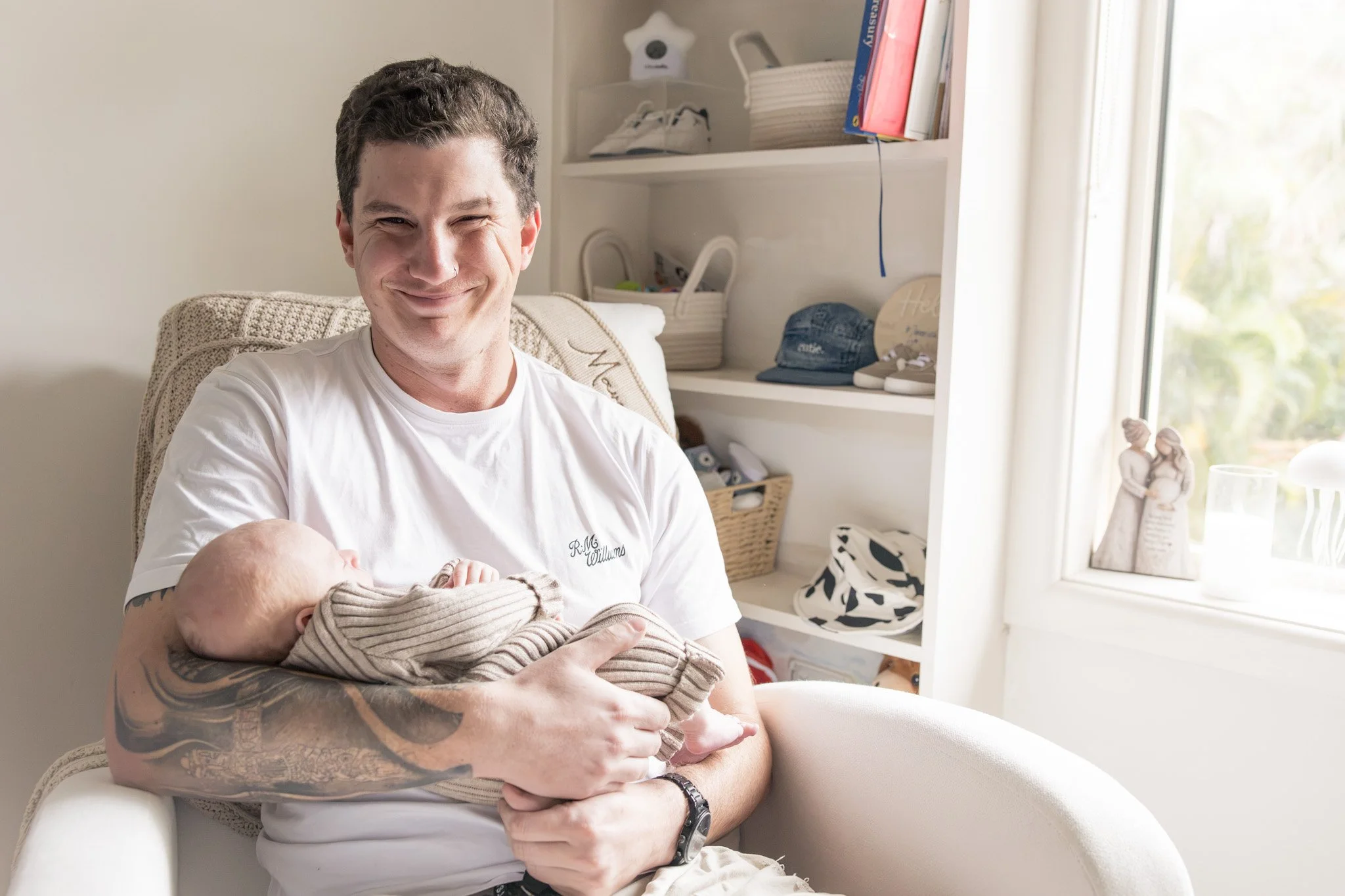 A man with tattoos holding a newborn baby in a cozy, well-lit room, sitting on a white chair with a smile.