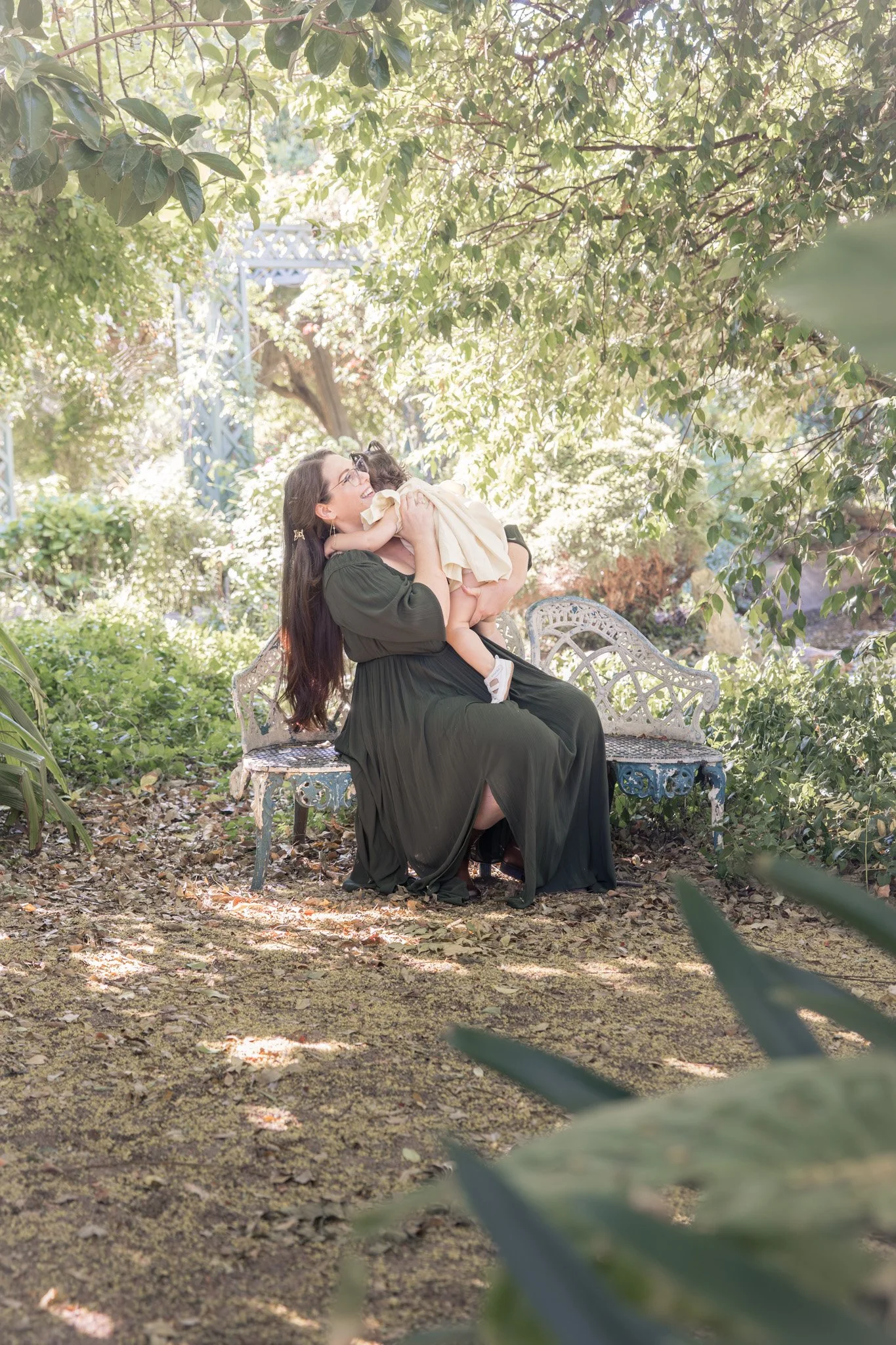 A woman with long dark hair, glasses, and a dark green dress, sitting on an ornate garden bench in a lush outdoor setting, holding and kissing a small child in a light yellow dress during a family photography session