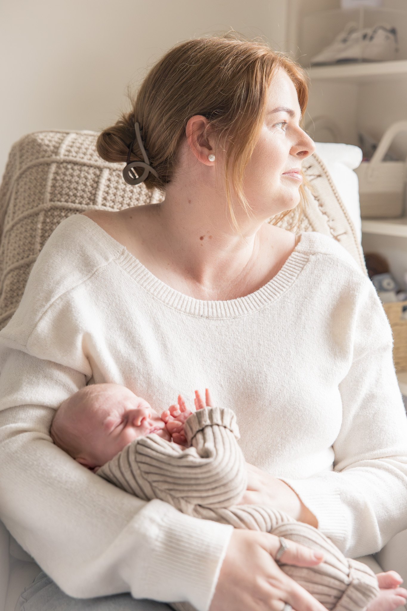 A woman with shoulder-length ginger hair and pearl earrings is sitting in a cozy beige armchair with a crochet pattern backrest. She is looking to the right with a gentle smile, wearing a white oversized sweater. She is holding a newborn baby wrapped